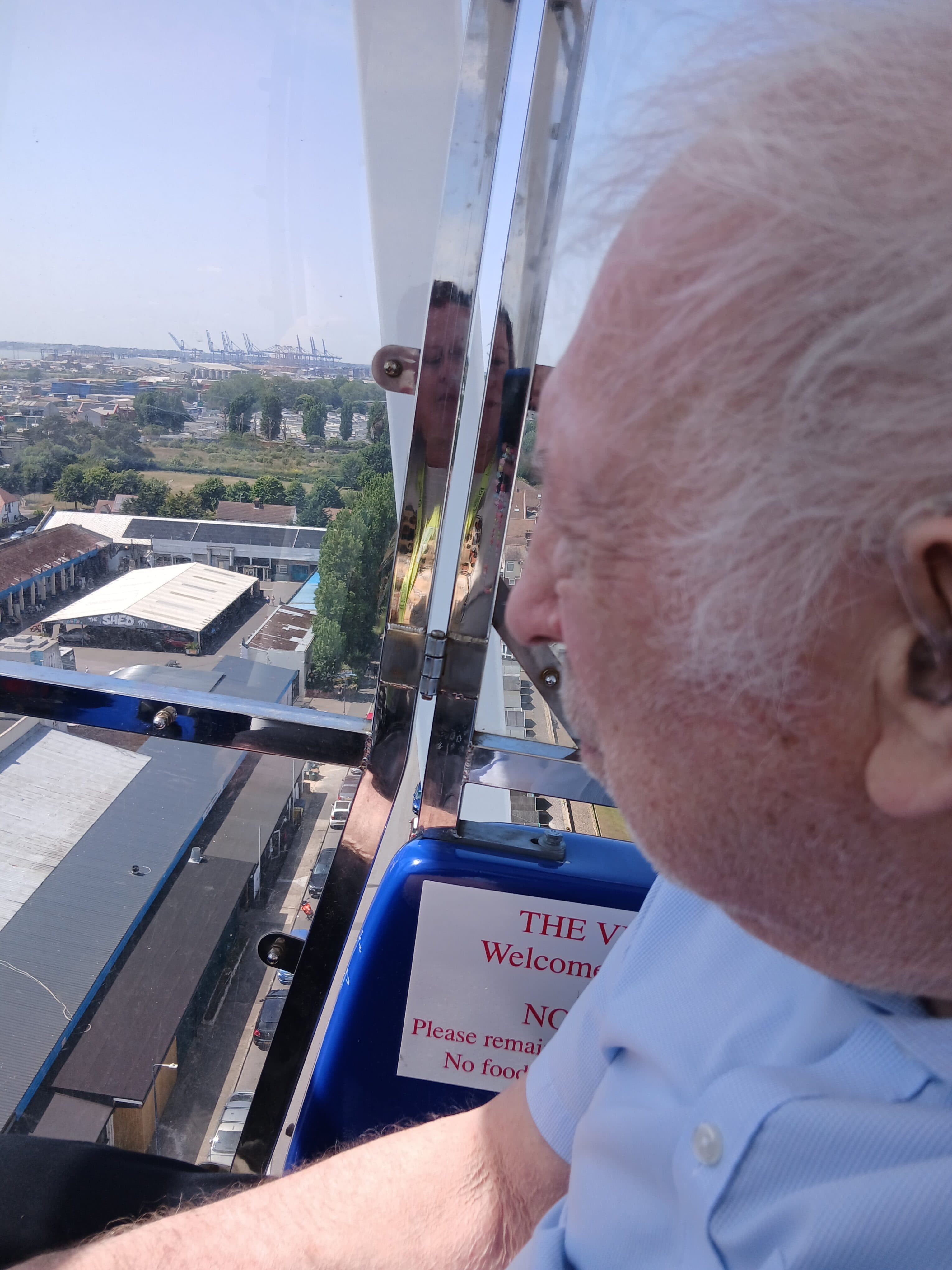 An elderly man looks out from a Ferris wheel gondola over a cityscape on a sunny day. - Home Instead