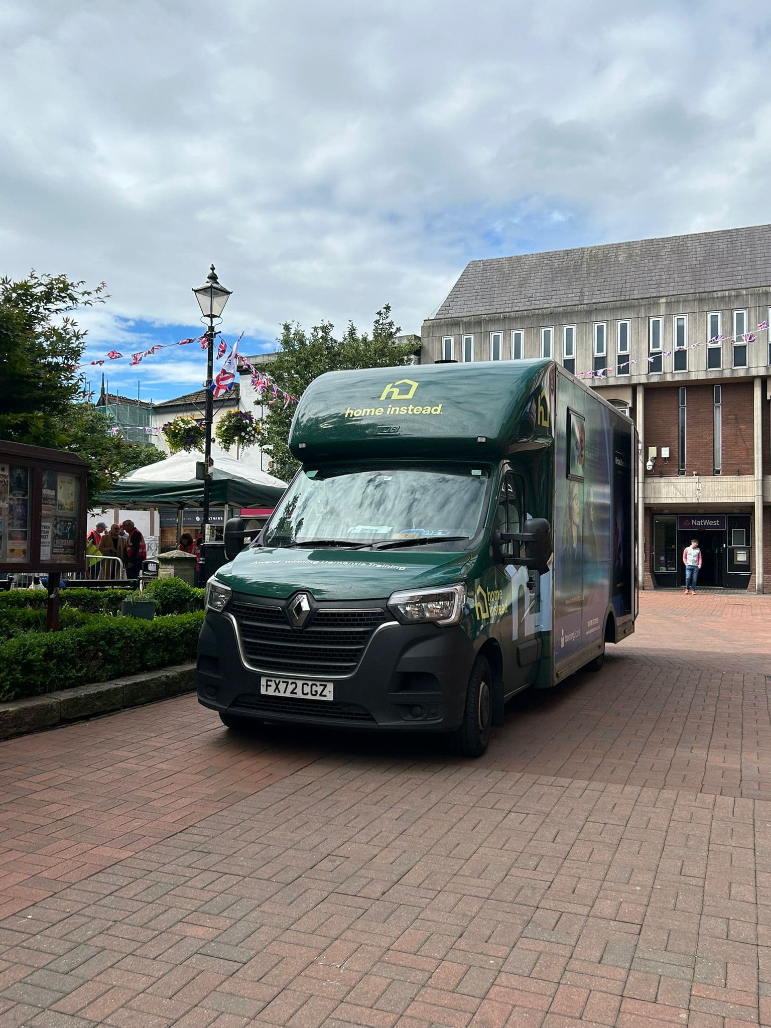 A green Home Instead van parked on a brick pathway in a town center with people and buildings nearby. - Home Instead