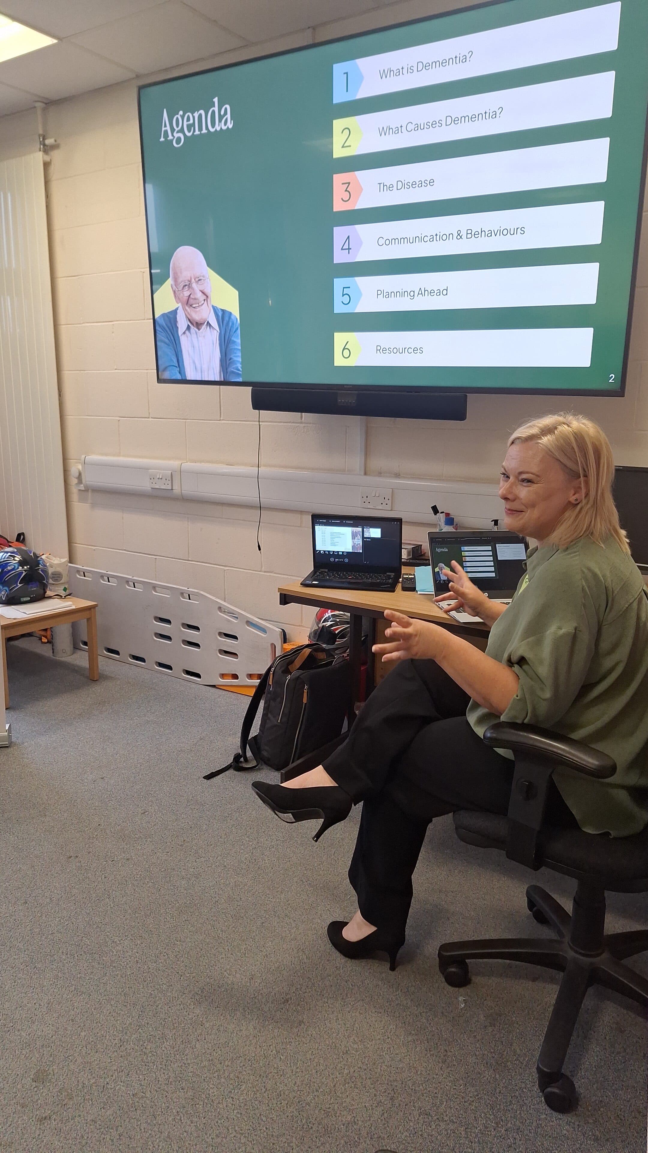 Woman leads a presentation on dementia; agenda is displayed on a large screen in a classroom setting. - Home Instead