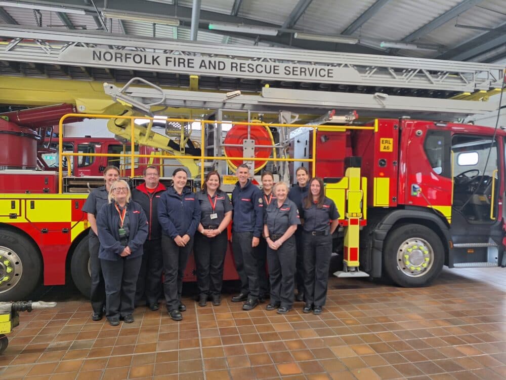 Twelve firefighters stand smiling in front of a Norfolk Fire and Rescue Service fire truck inside a station. - Home Instead