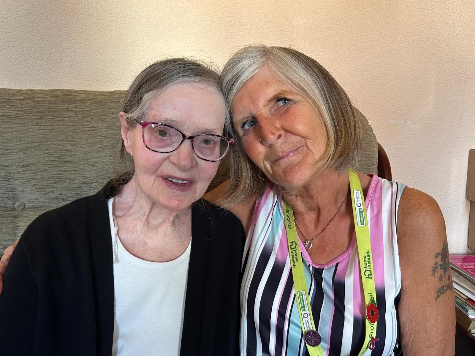 Two older women sit close together, smiling warmly at the camera in a cozy indoor setting. - Home Instead