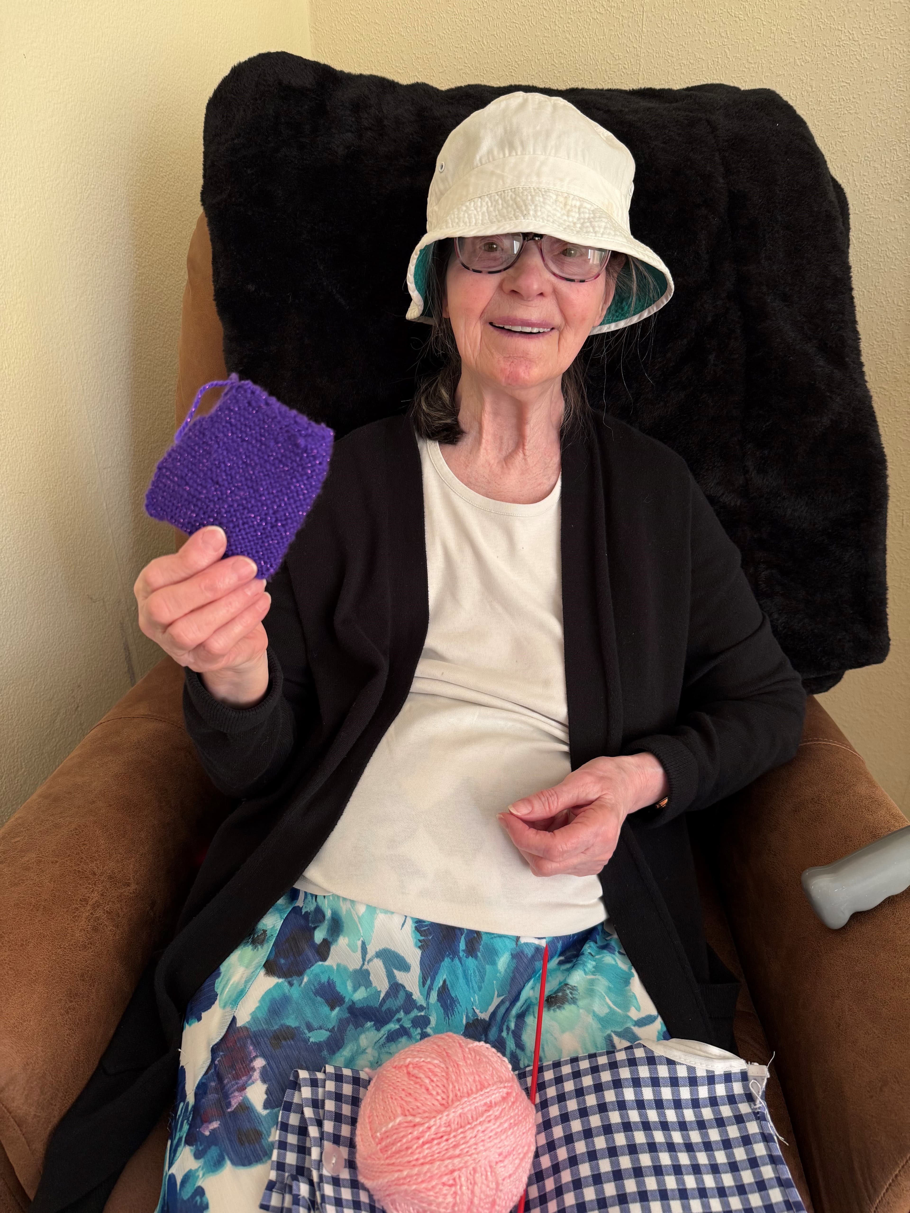 Smiling elderly woman in a hat holds up purple knitting, seated with yarn and fabric on her lap. - Home Instead