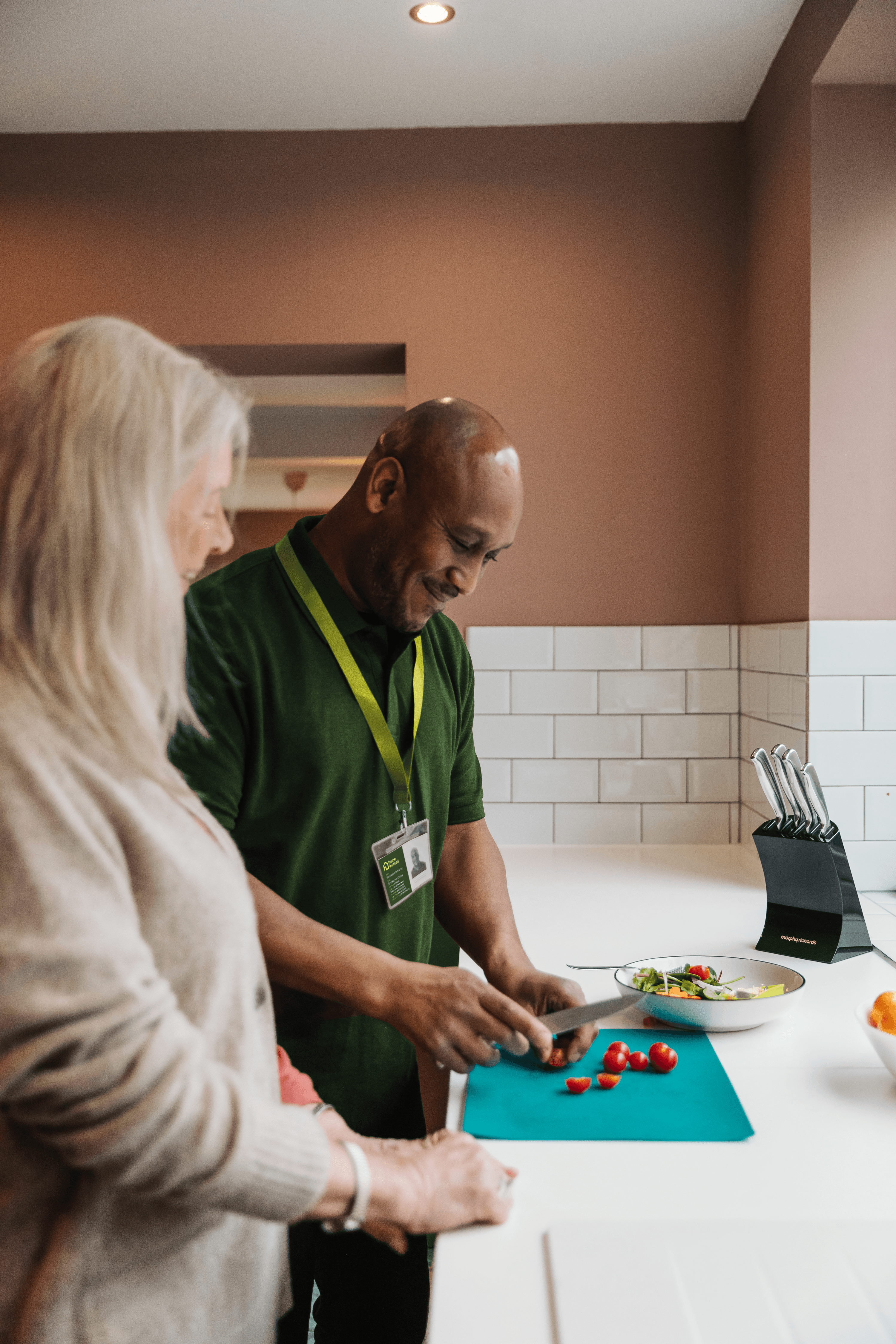 A man in a green top helps an elderly woman in a kitchen cut vegetables.