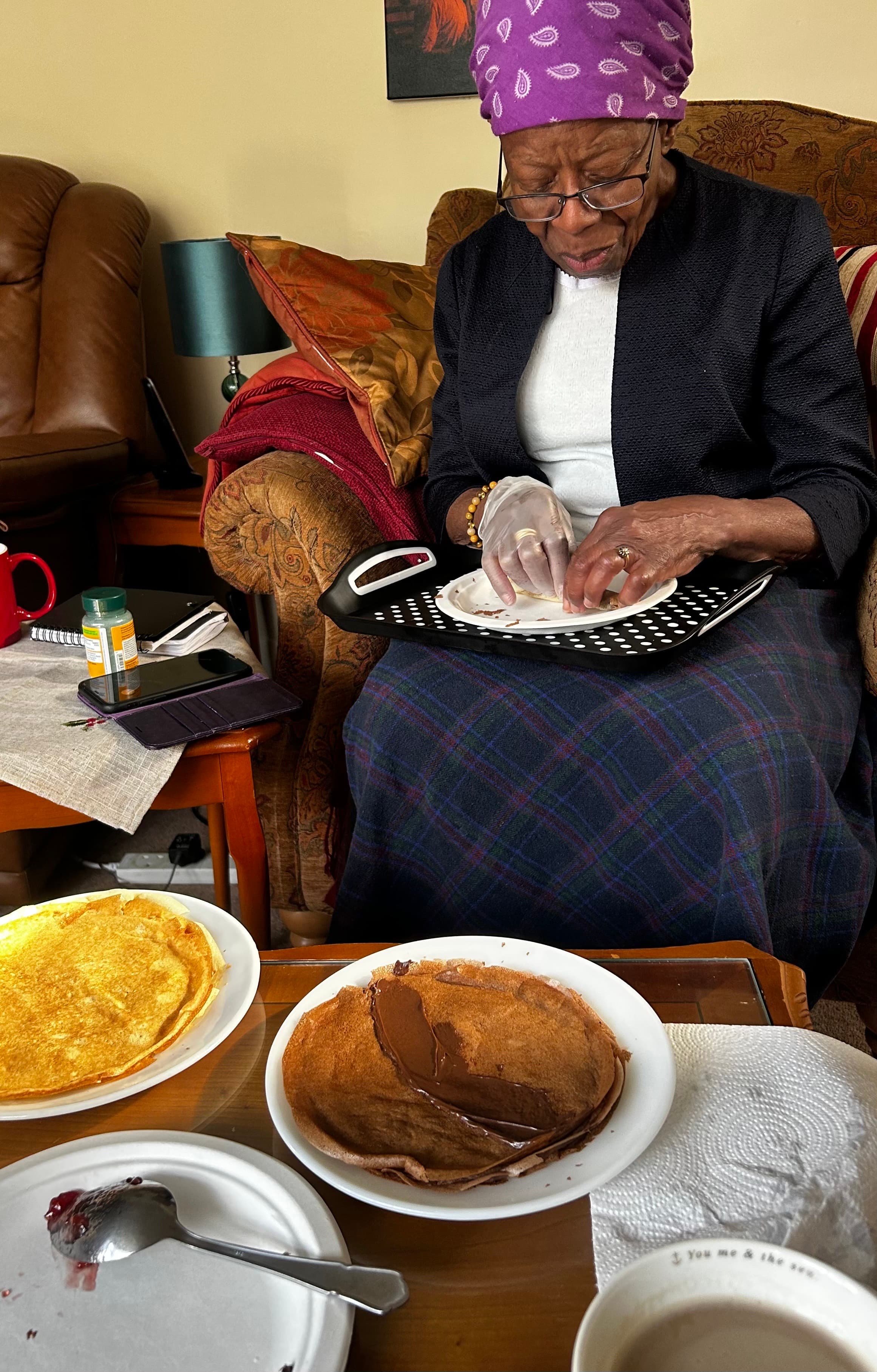 Elderly woman in purple headscarf prepares food on her lap; pancakes, coffee, and jam are on the table. - Home Instead