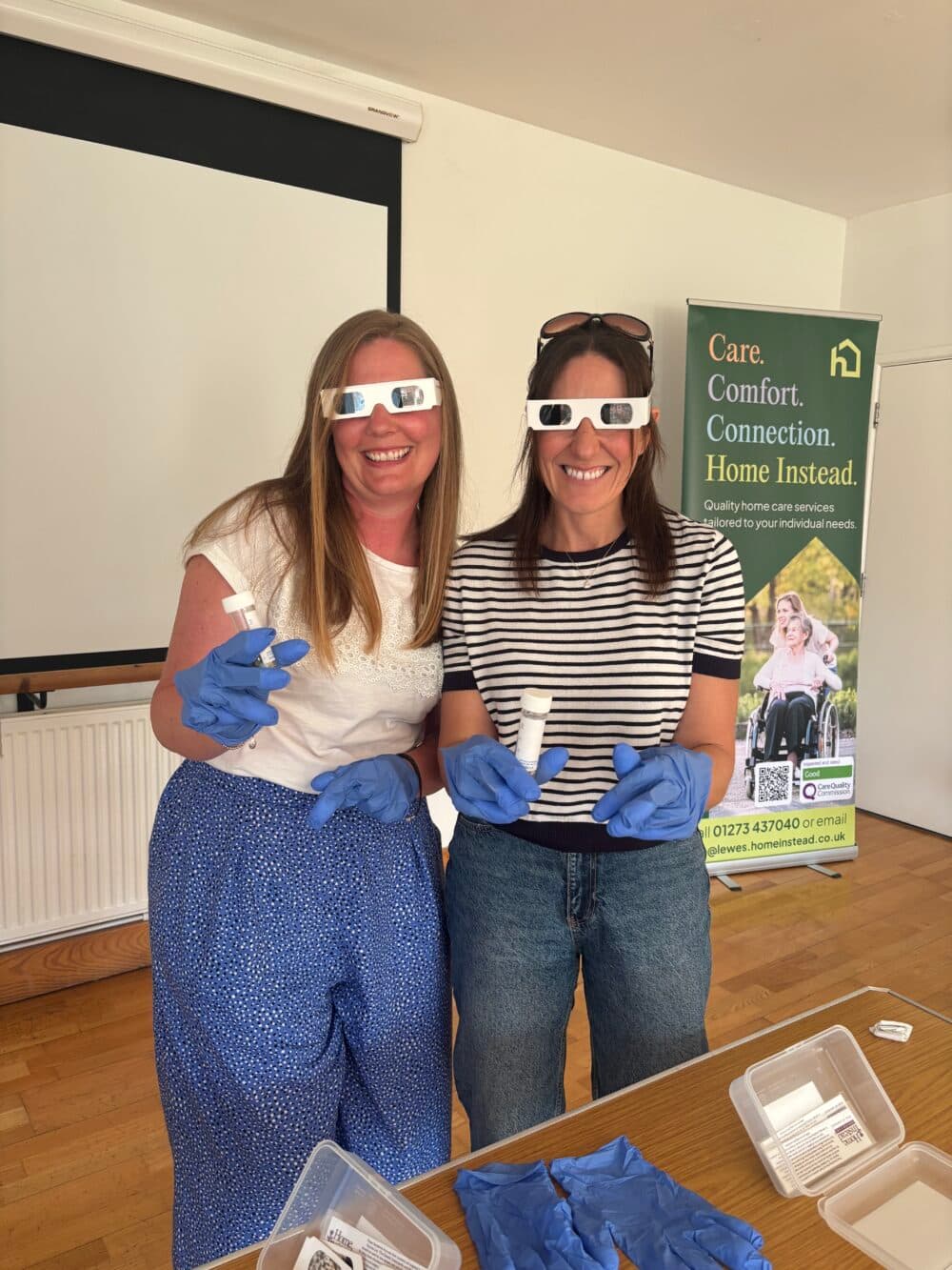 Two women wearing gloves and 3D glasses smile, holding objects, standing by a display table and a care services banner. - Home Instead