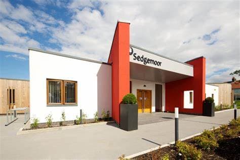 Modern building with white and red exterior, labeled "Sedgemoor," under a blue sky with scattered clouds. - Home Instead