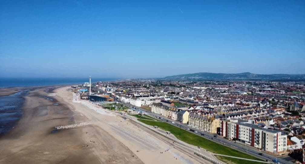 Aerial view of a seaside town with sandy beach, promenade, and buildings under a clear blue sky. - Home Instead