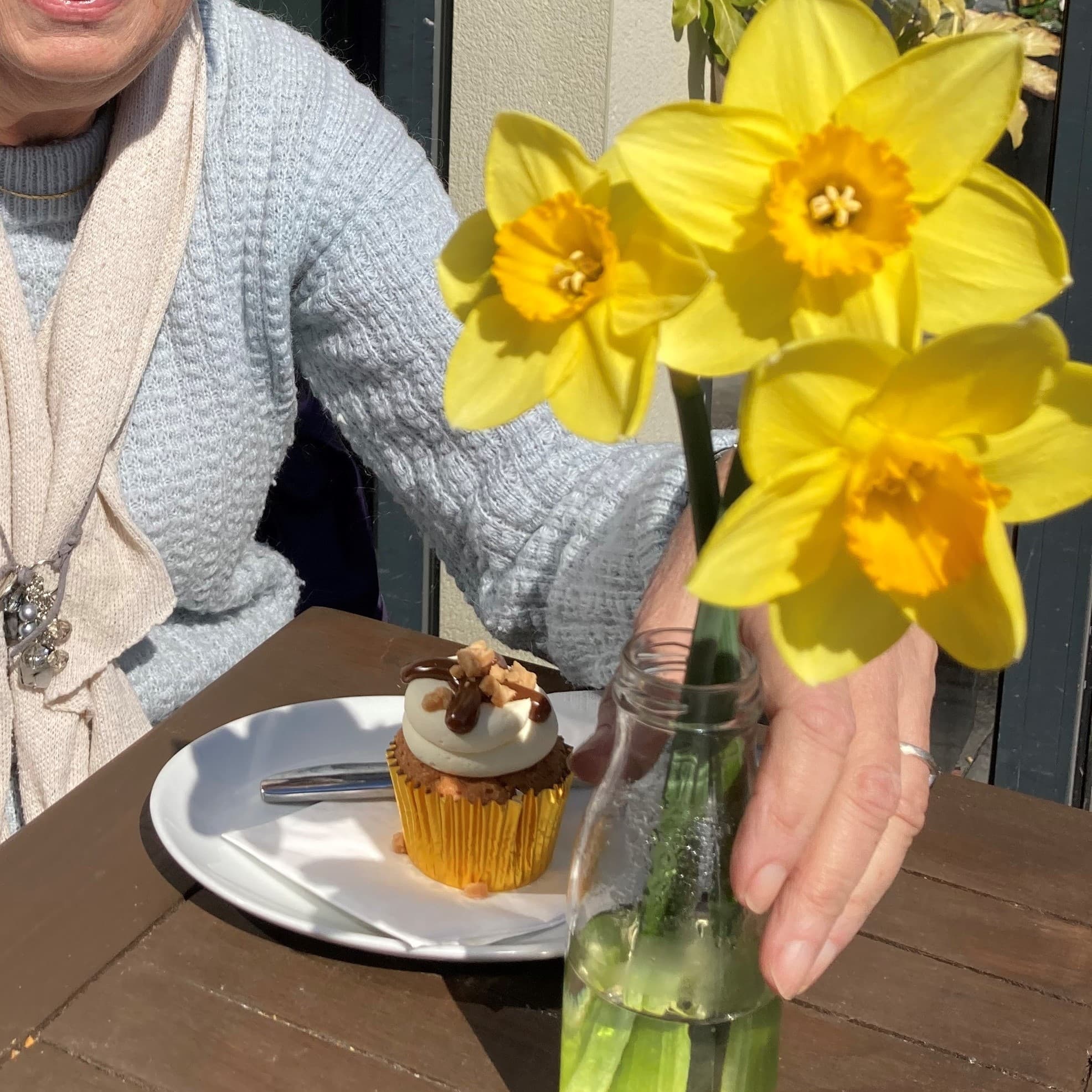 A person in a gray sweater sits at a table with a cupcake and yellow daffodils in a glass vase. - Home Instead