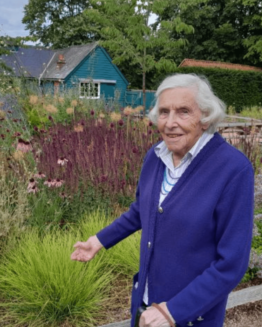 An elderly woman in a blue coat stands in a colorful garden, smiling at the camera. - Home Instead