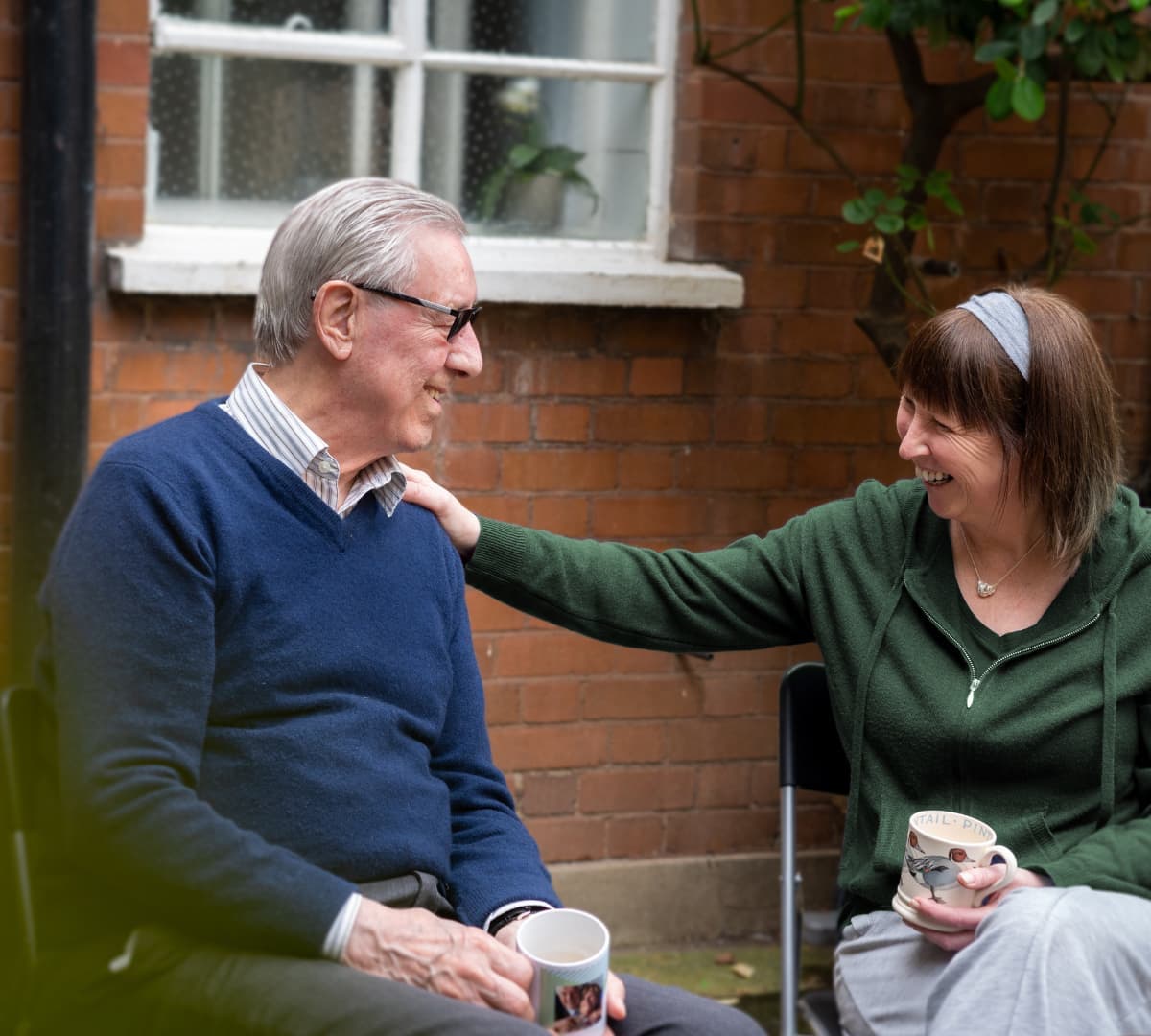 An older man and a woman smiling and talking while holding mugs, sitting outside by a brick wall. - Home Instead