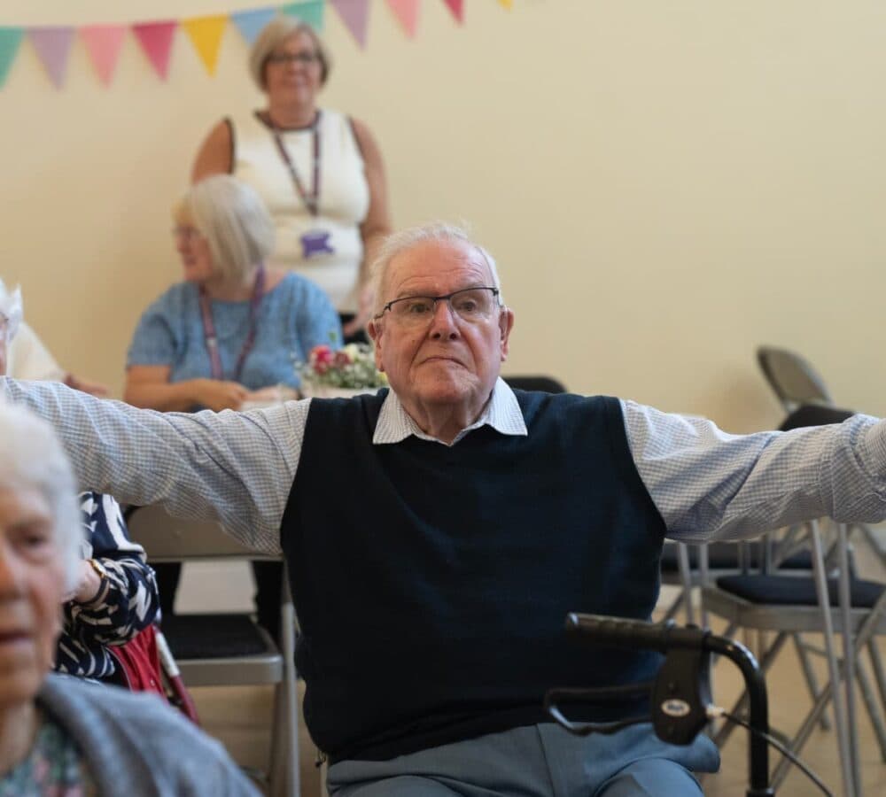 An older male adult with white hair and wearing eyeglasses and blue vest smiling and happy while doing some chair exercise inside the room with other older adults