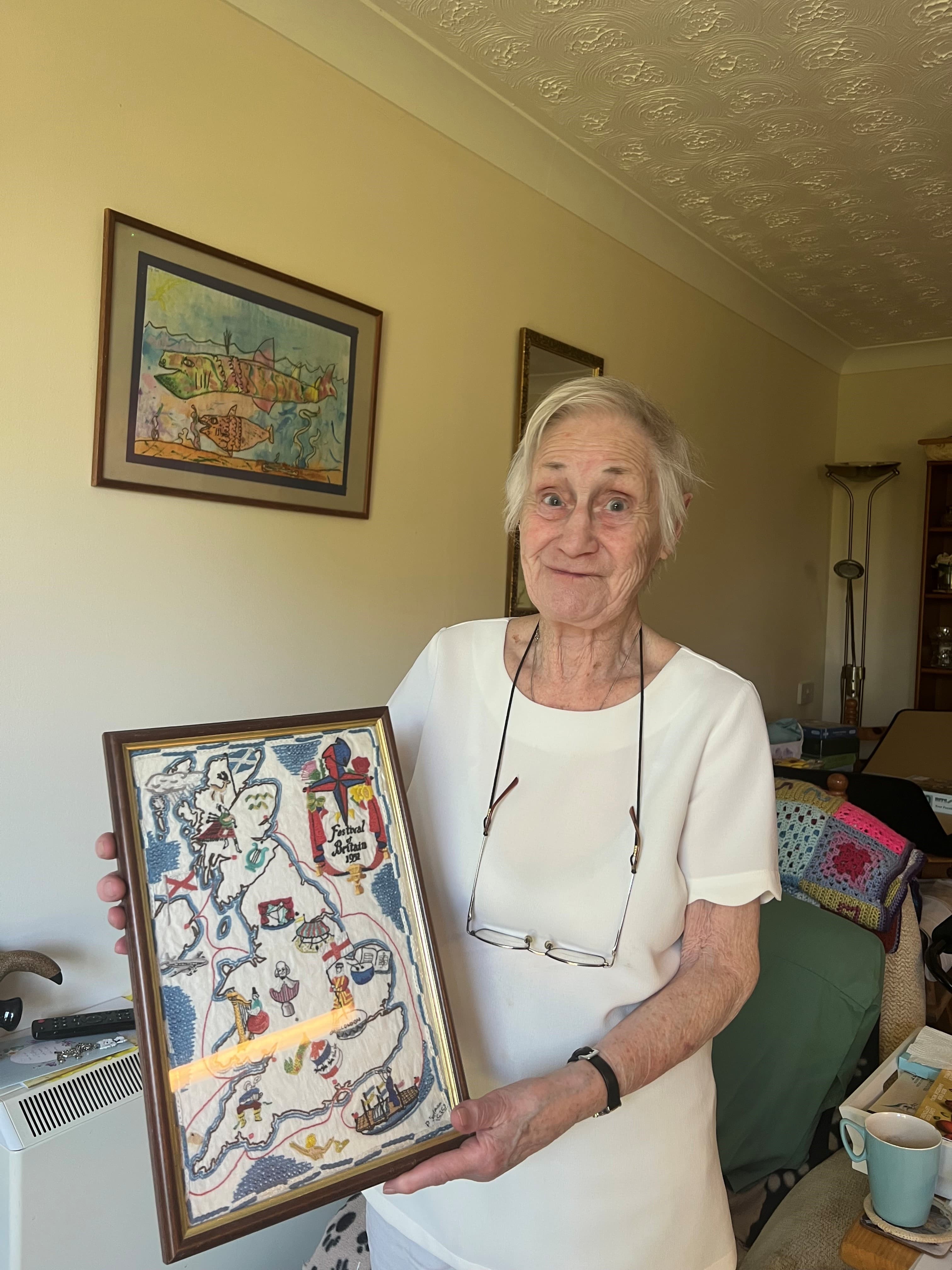 Elderly woman smiling and holding a framed embroidered map in a cozy living room. - Home Instead