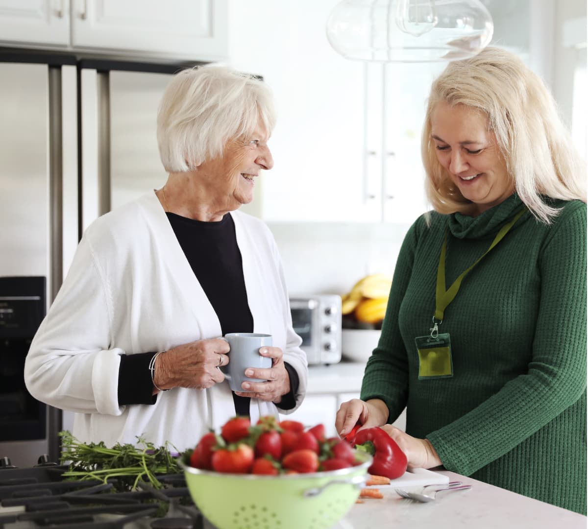 An older woman and a younger woman smile and prepare food together in a bright kitchen. - Home Instead