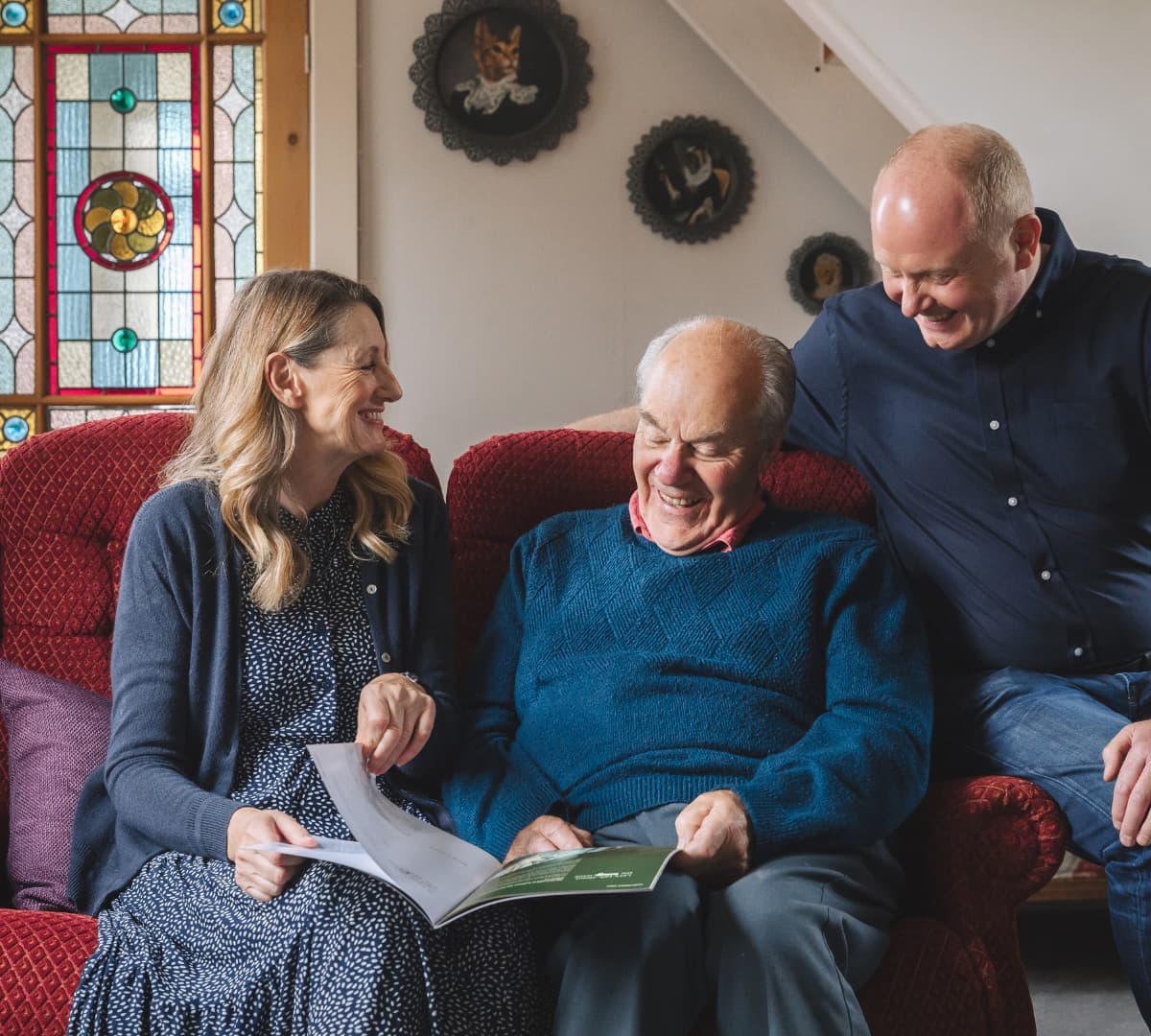 An older male adult sitting on a couch while reading a magazine with his daughter and son inside the house, all are wearing blue