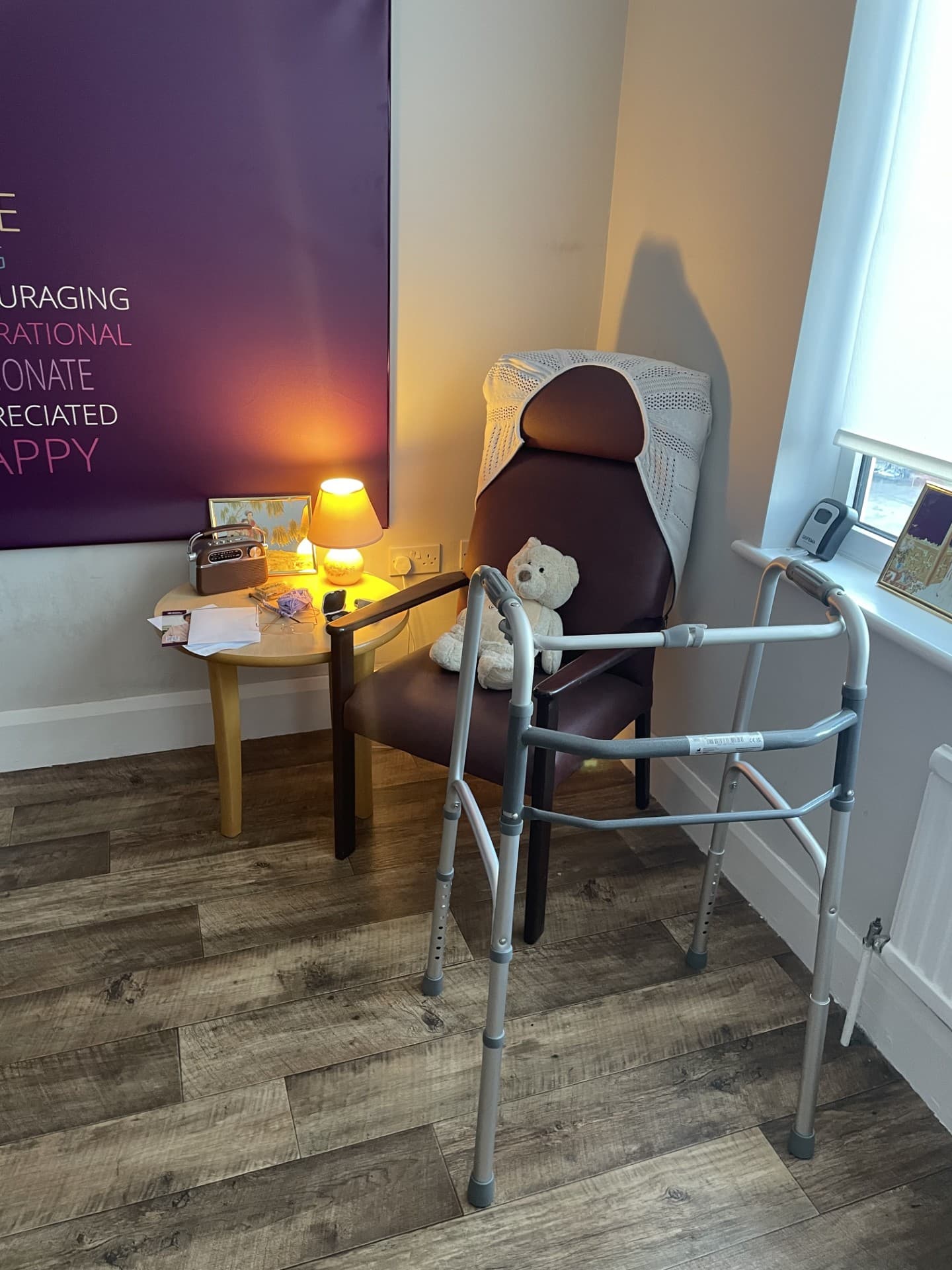 A chair with a teddy bear, a walker in front, and a small table with a lamp and items beside it. - Home Instead