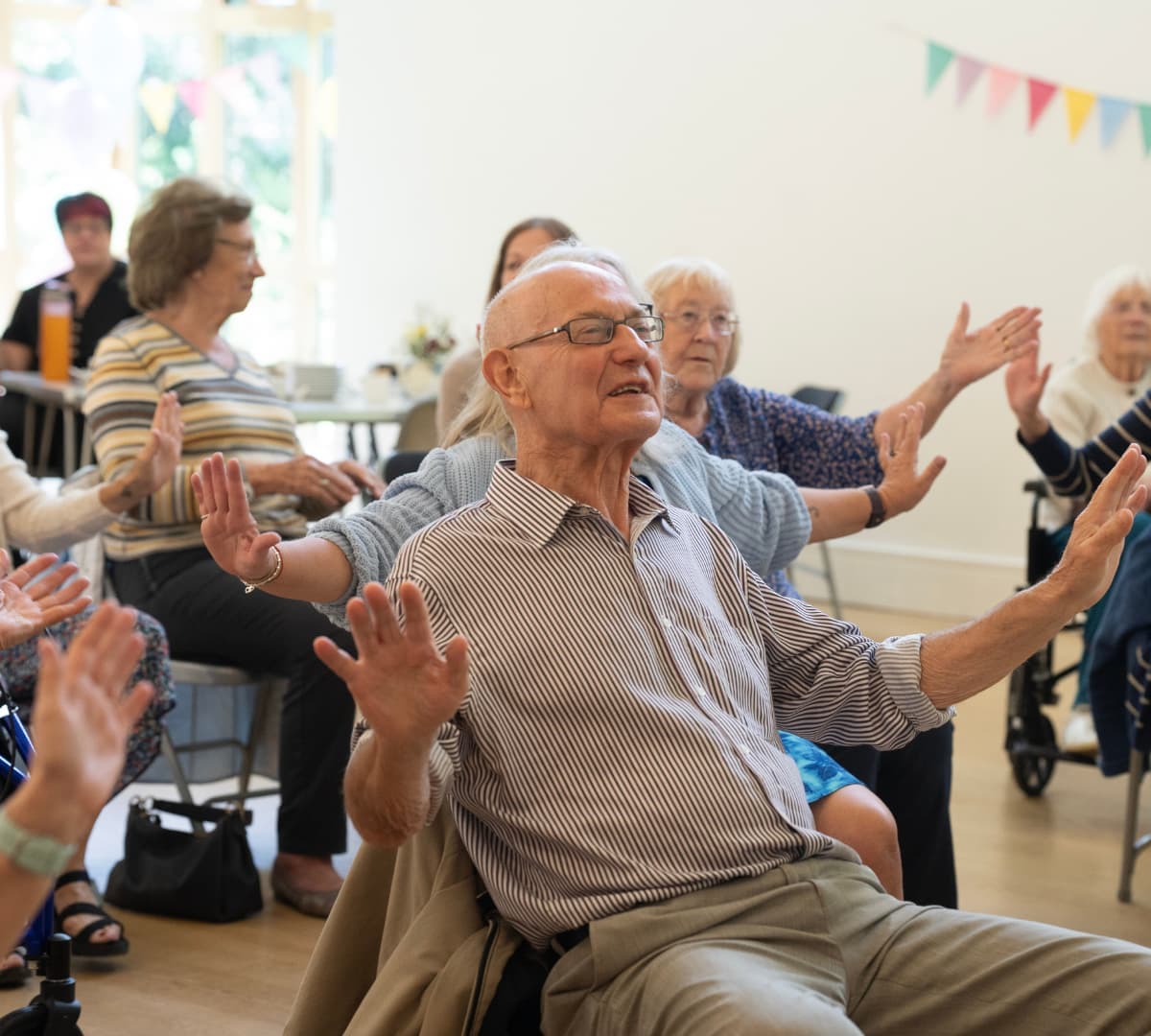 An older male adult with white hair and wearing eyeglasses smiling and happy while doing some chair exercise inside the room with other older adults