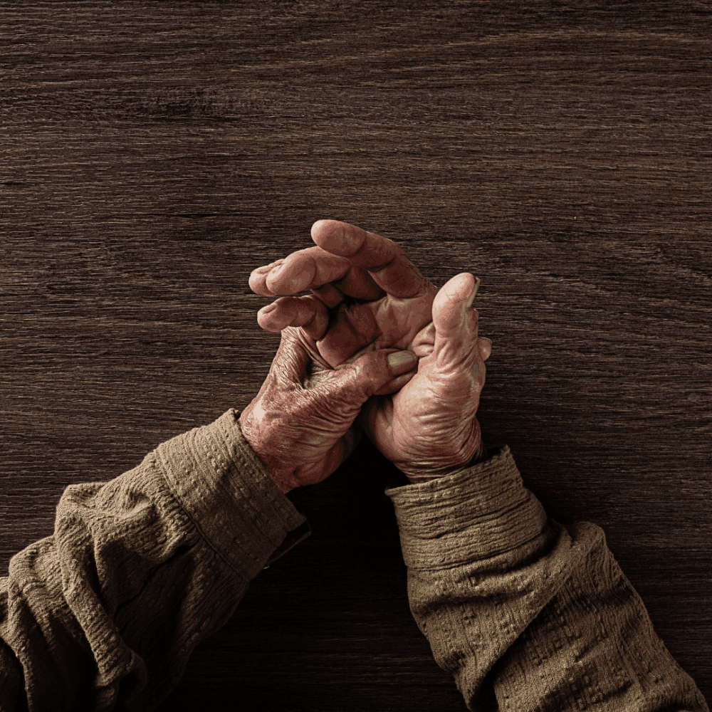 Elderly hands clasped together on a wooden surface, wearing a textured brown long-sleeve shirt. - Home Instead