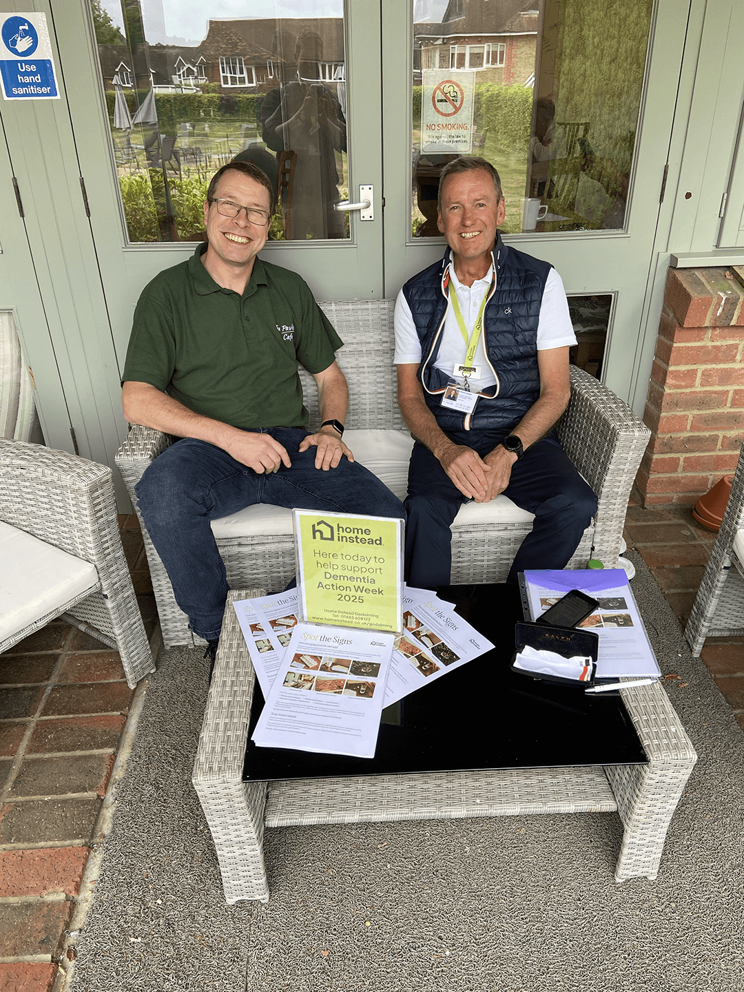 Two men sitting outside a cafe. On the left man in glasses wearing green polo shirt and jeans. Man on the right wearing a white shirt and blue sleeveless jacket and blue trousers with a lanyard on from Home Instead.
