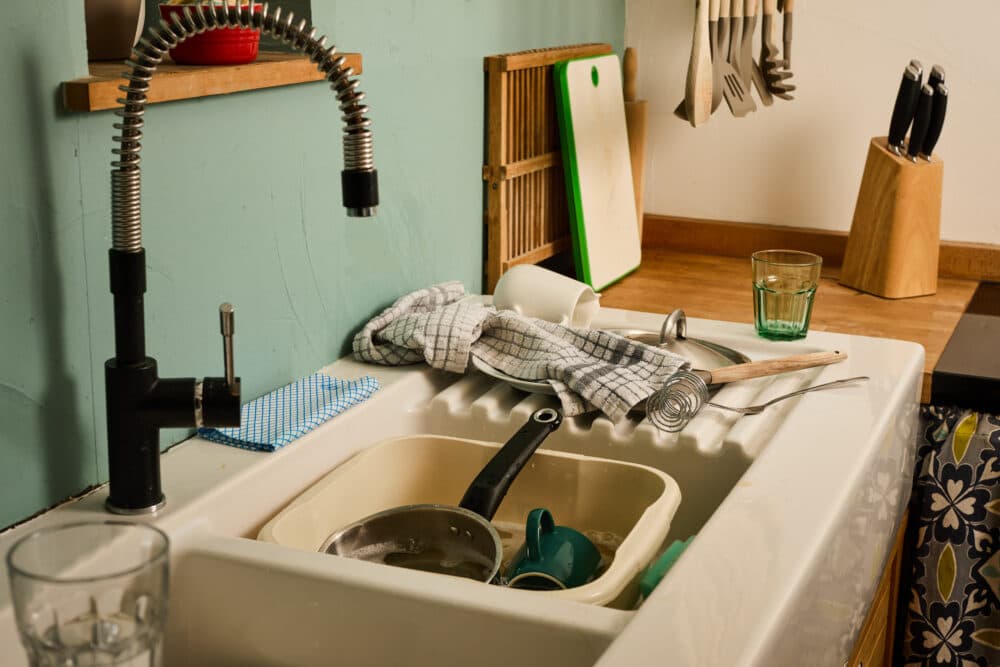 A kitchen sink filled with dirty dishes, utensils, and cups, with a towel draped over the edge. - Home Instead