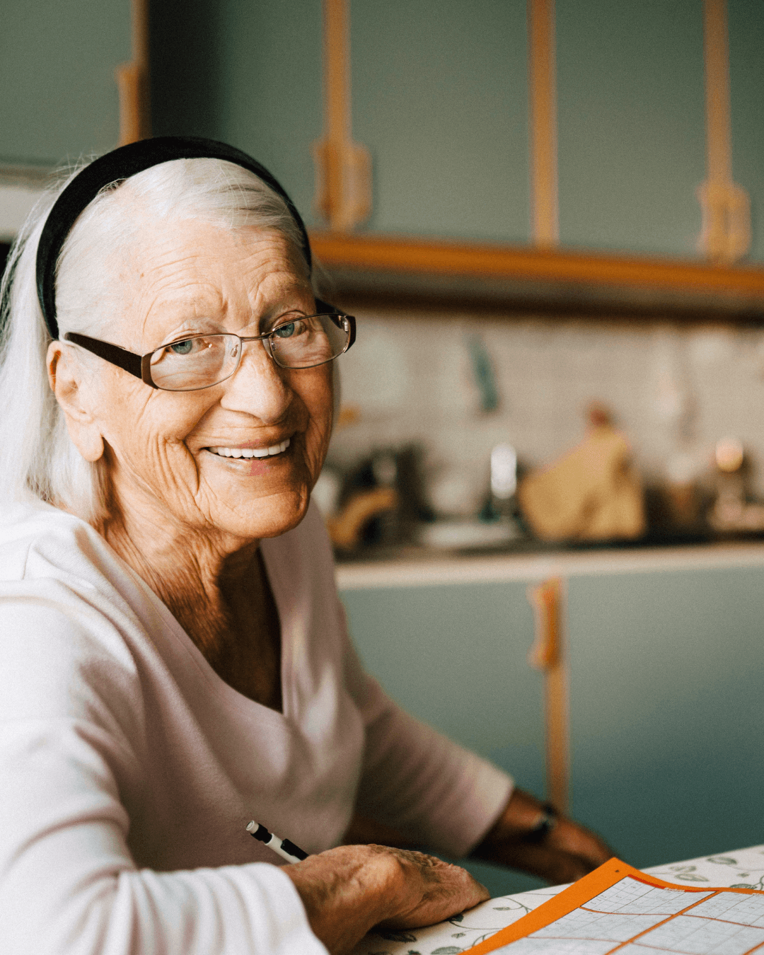 Smiling elderly woman with glasses, seated at a table, holding a pencil in a cozy kitchen. - Home Instead