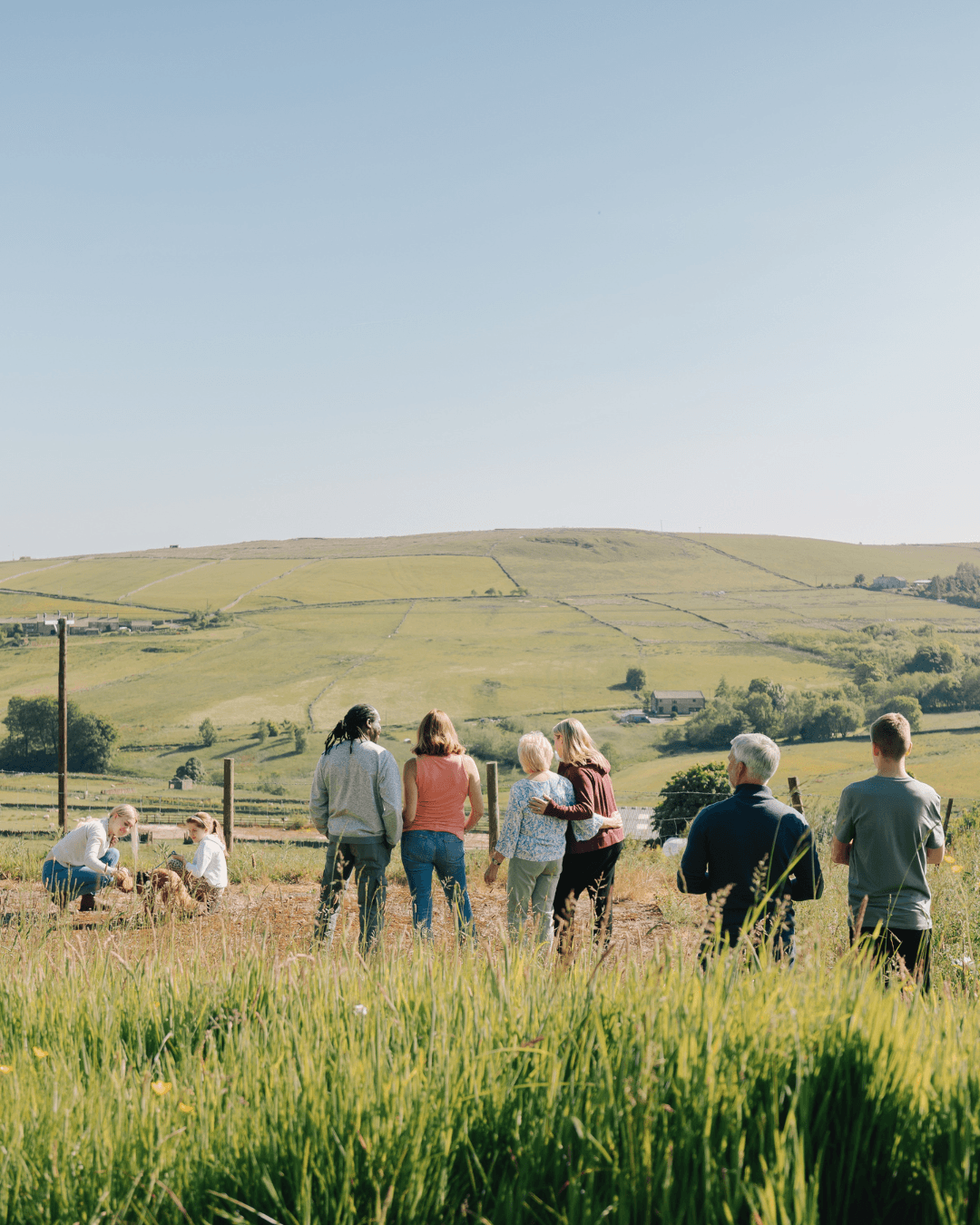 A group of people stand and sit in a grassy field, looking out at rolling green hills under a clear sky. - Home Instead