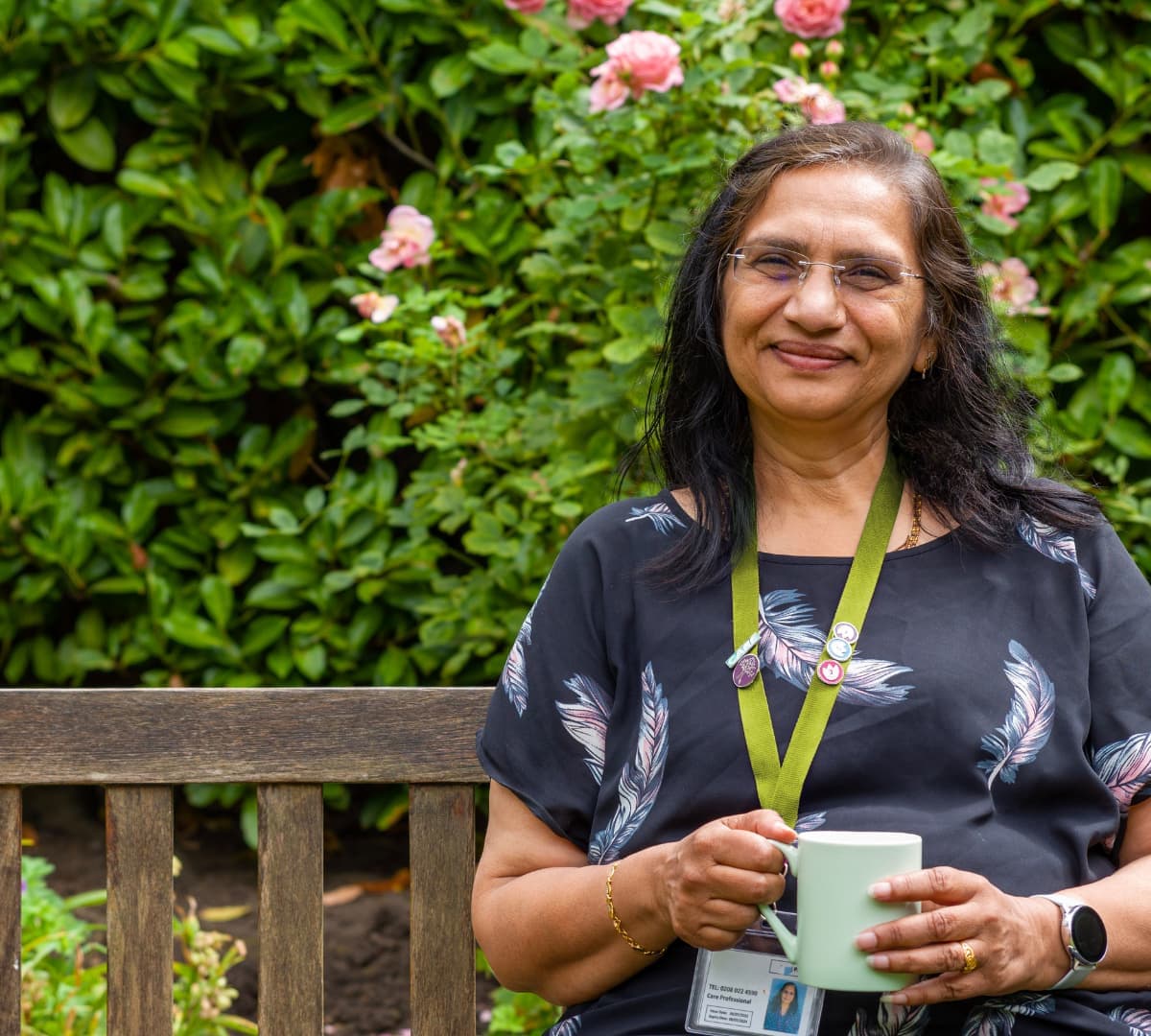A woman with long black hair happy and smiling while sitting on a bench and holding a cup of coffee in the garden