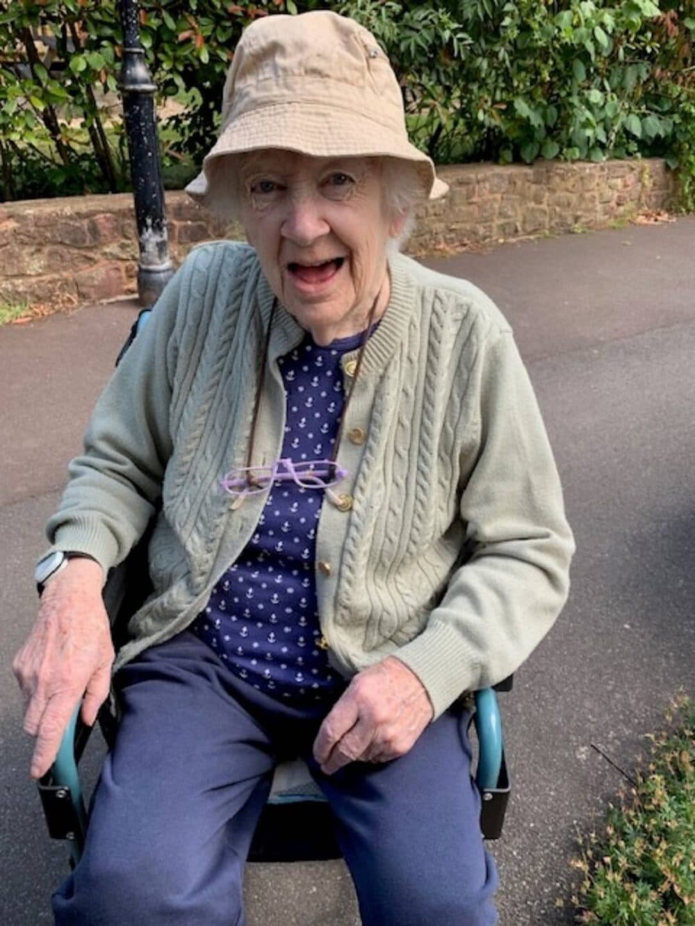 Smiling elderly woman in a sun hat and cardigan sitting outside in a wheelchair on a paved path. - Home Instead