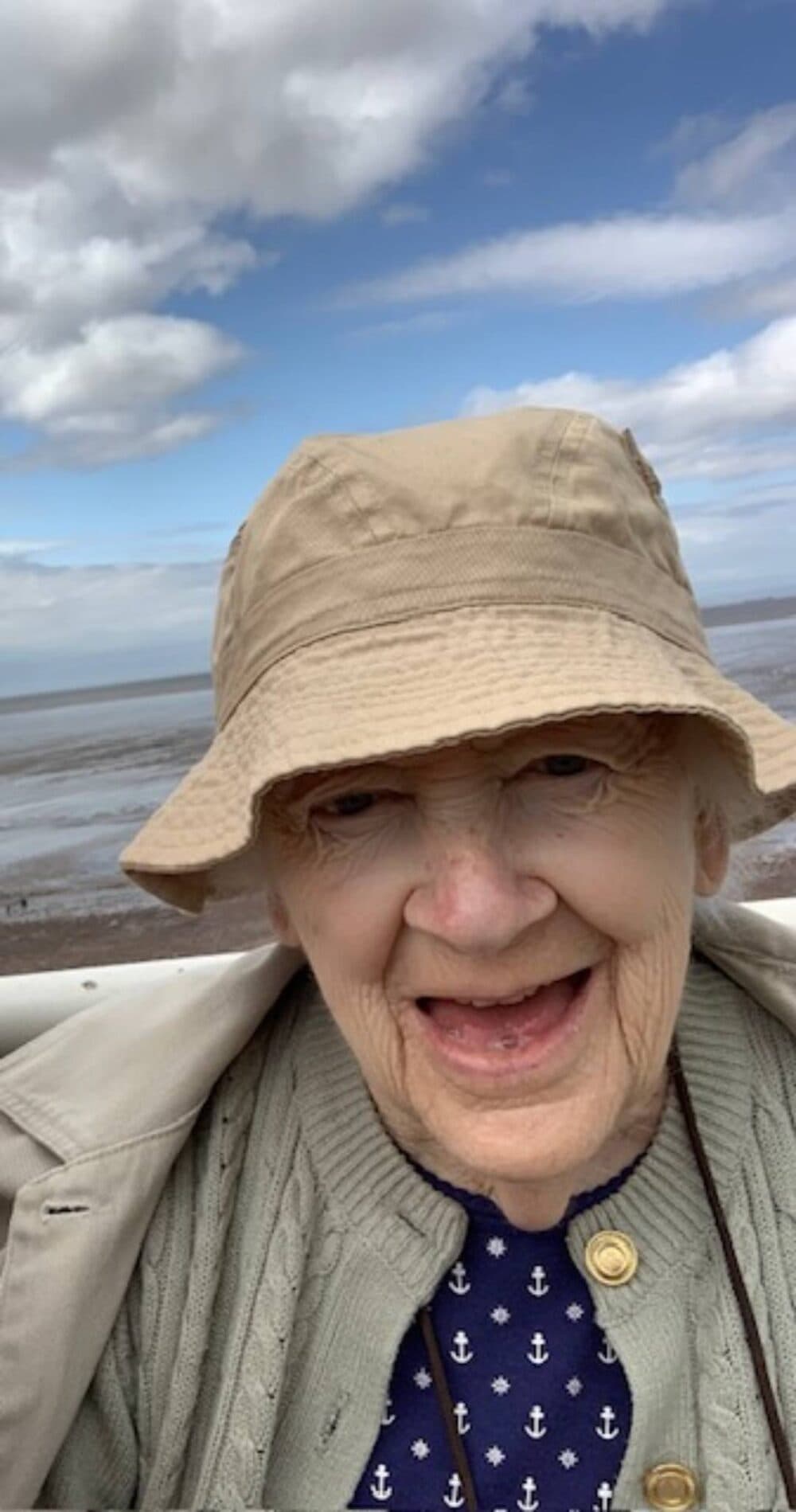 Smiling elderly woman in a beige hat and cardigan, outdoors by the beach under a partly cloudy sky. - Home Instead