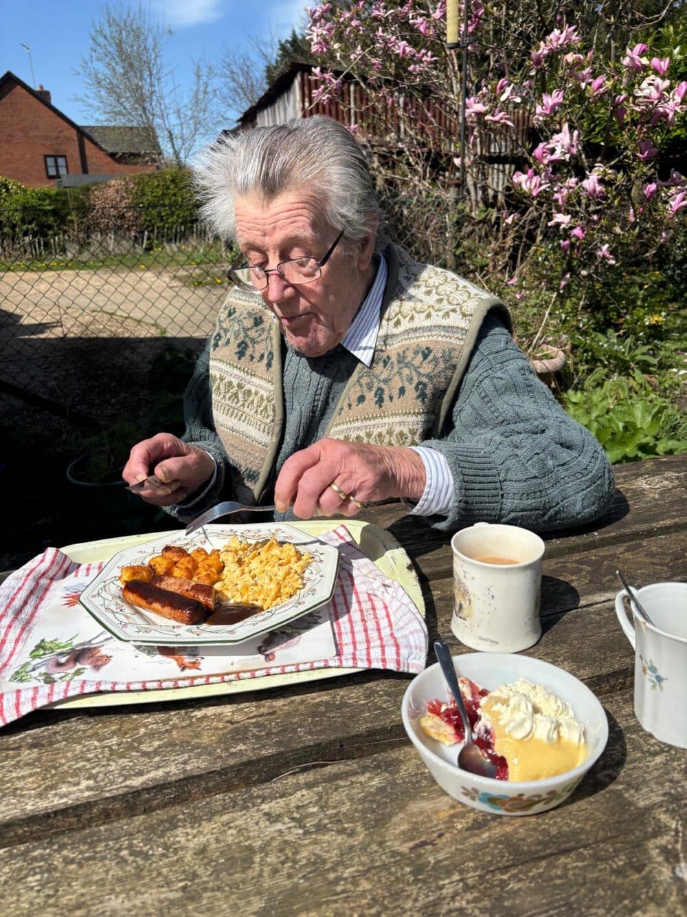 Elderly man eating breakfast outdoors at a wooden table with tea and dessert on a sunny day. - Home Instead