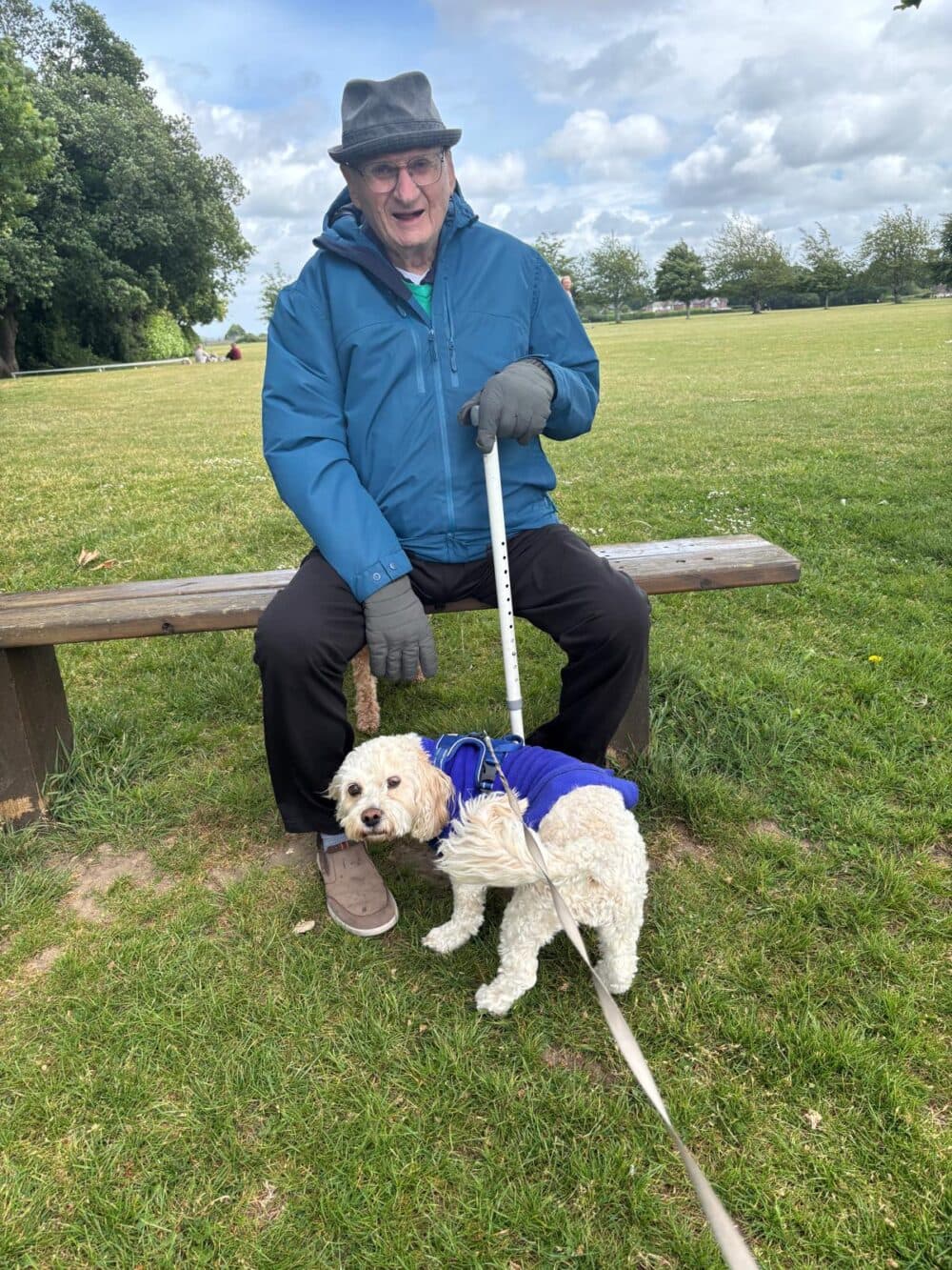 An older man sits on a bench holding a white dog on a leash in a grassy park. - Home Instead