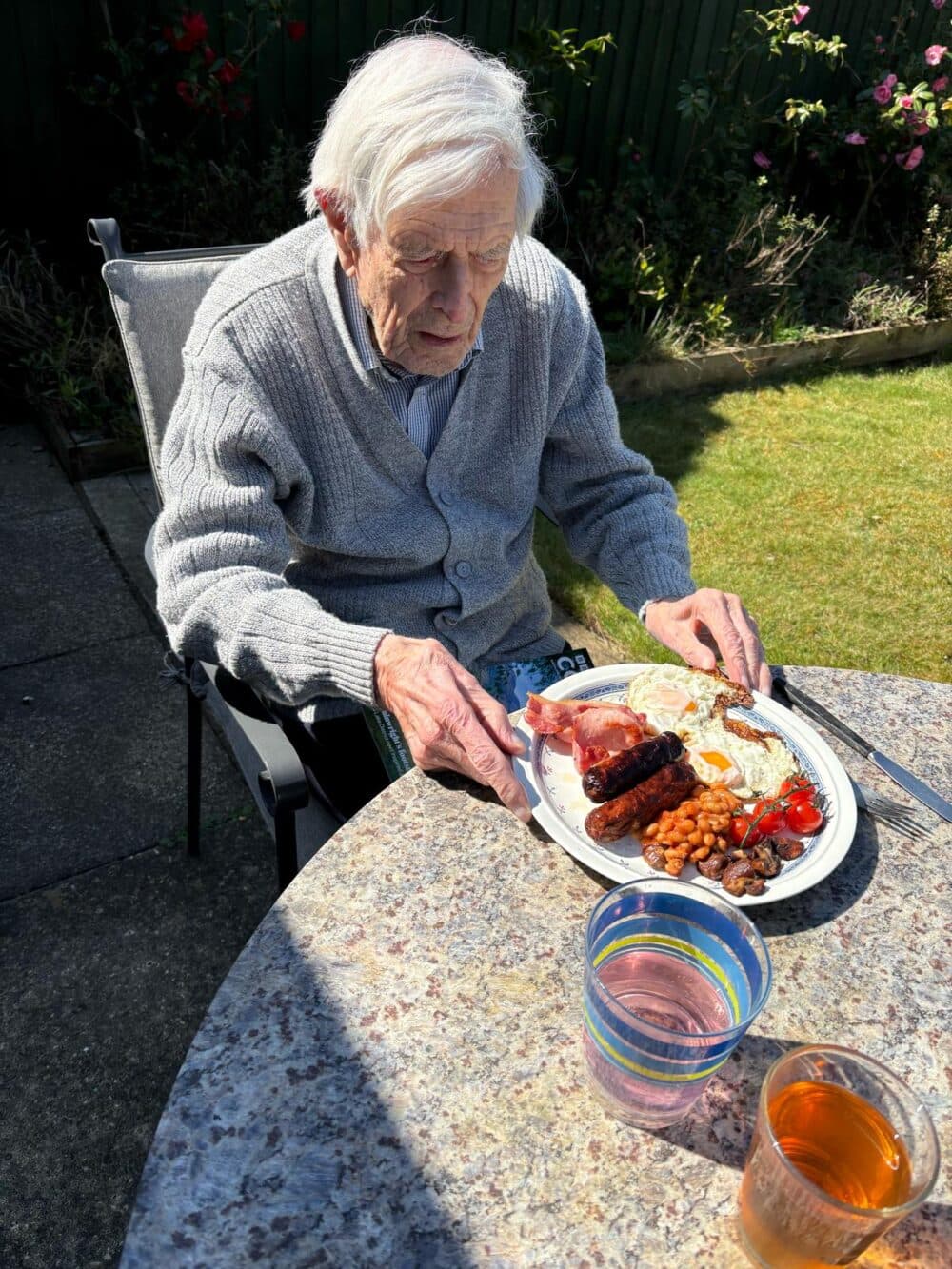 Elderly man in a grey cardigan eating a full English breakfast at a garden table in the sunshine. - Home Instead