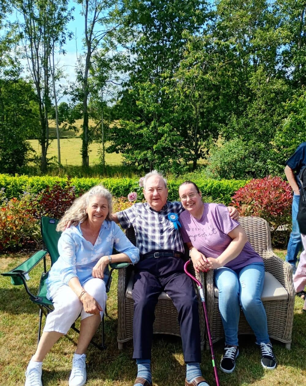 Three people sit smiling together outdoors on a sunny day, surrounded by green trees and bushes. - Home Instead