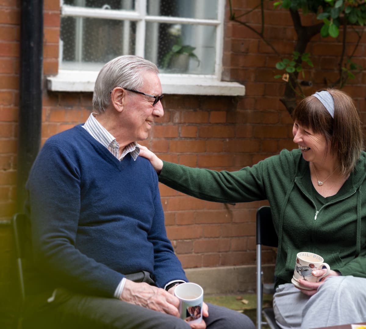 An older male adult with grey hair and wearing blue eyeglasses and ble sweatshirt chatting with his younger female carer with short hair and wearing reen bot happy and smiling outside the house