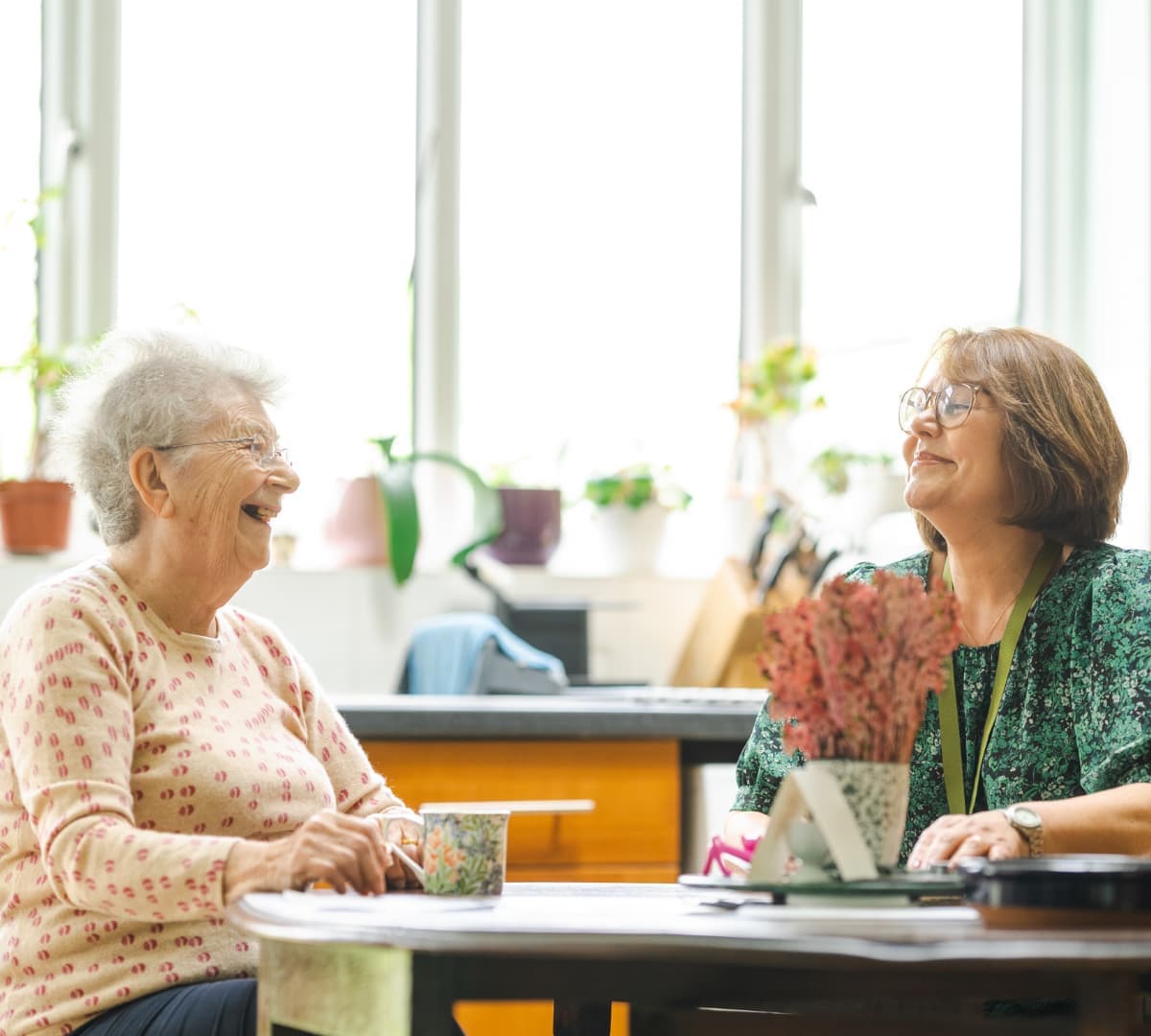An older female adult with grey hair chatting with her younger female carer wearing green while sitting on a chair and with coffee's on the table inside the kitchen