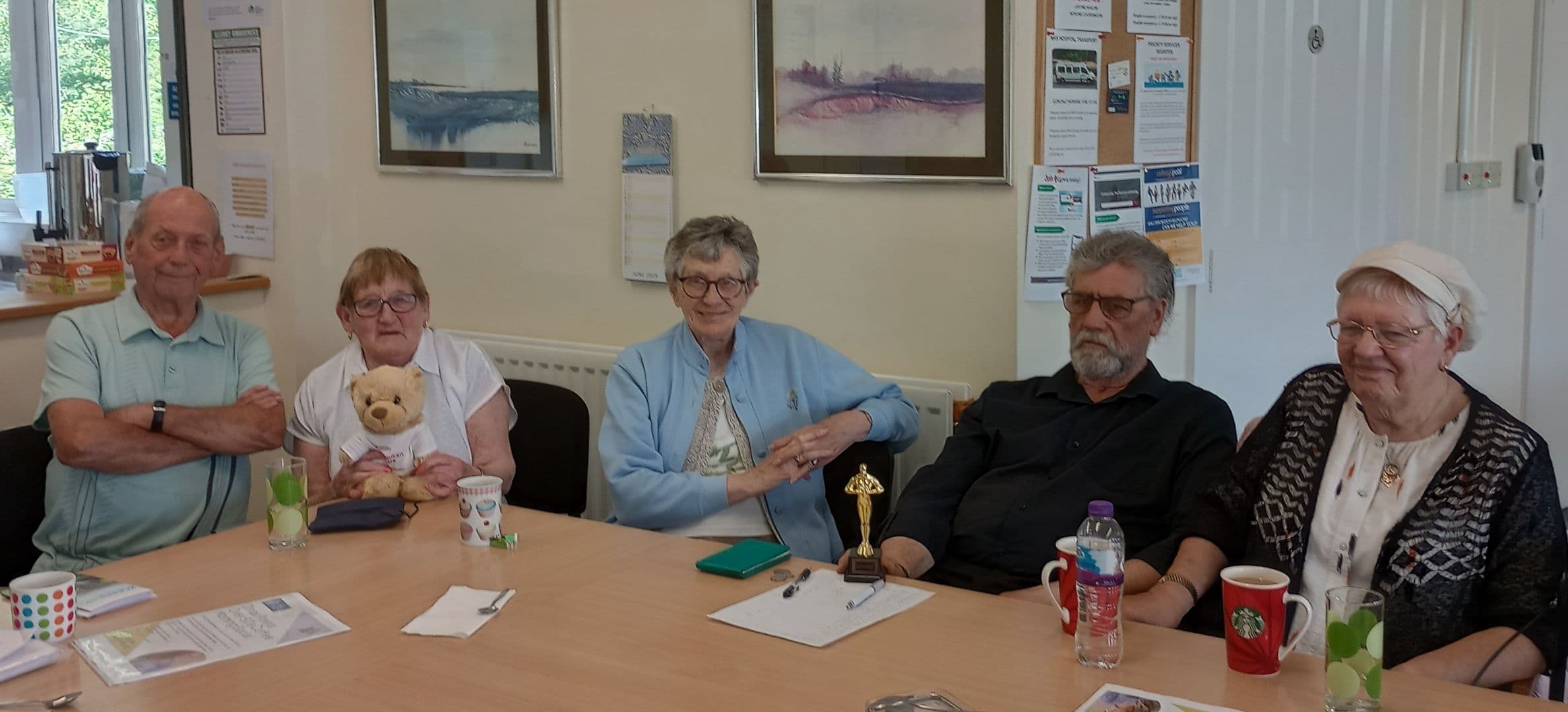 Five seniors sit at a table with drinks and notes; one holds a teddy bear, another an award. - Home Instead