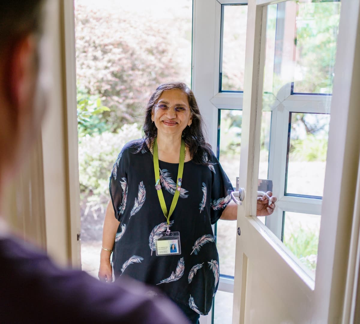 Smiling woman with a lanyard opens a door to greet a person standing outside. - Home Instead