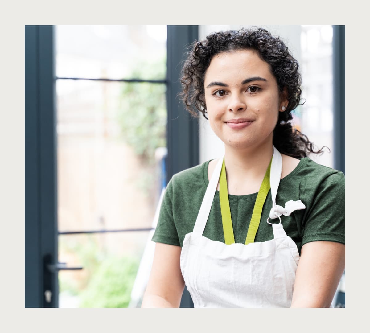 Woman with black curly hair and wearing green top with Apron and standing inside the kitchen