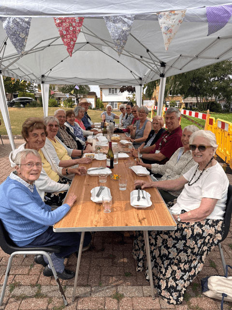 A group of older adults sit around a long table outdoors under a canopy, smiling and enjoying a meal together. - Home Instead