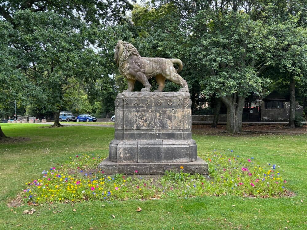 Stone lion statue on a pedestal surrounded by grass, trees, and colorful flowers in a park. - Home Instead