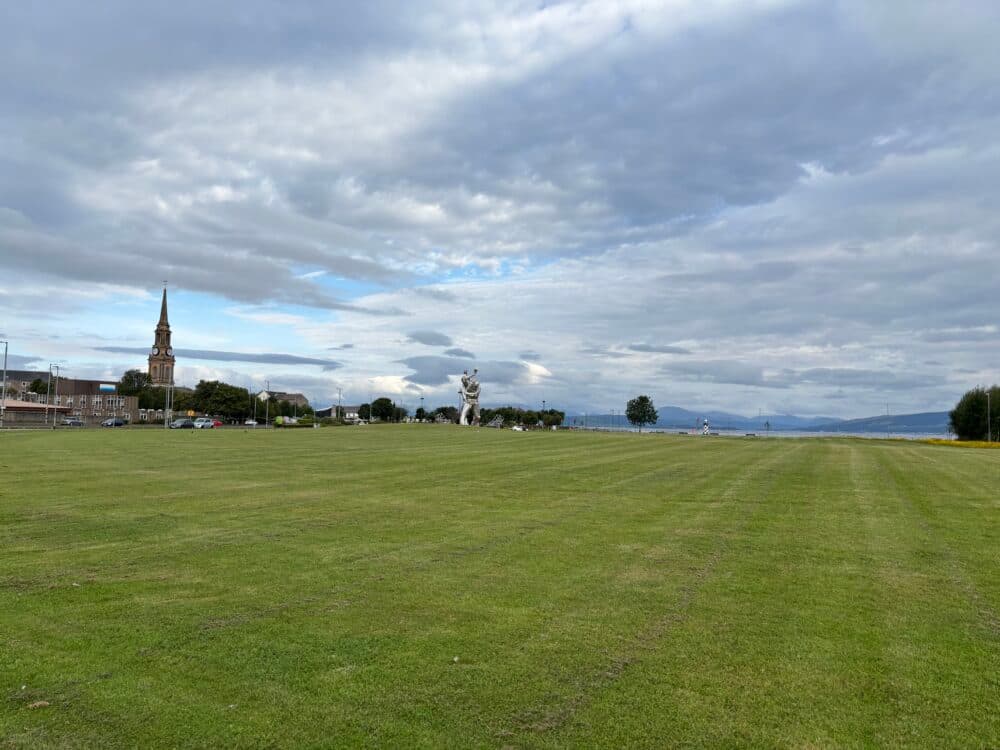 Wide grassy field with a church, statue, trees, and distant hills under a cloudy sky. - Home Instead