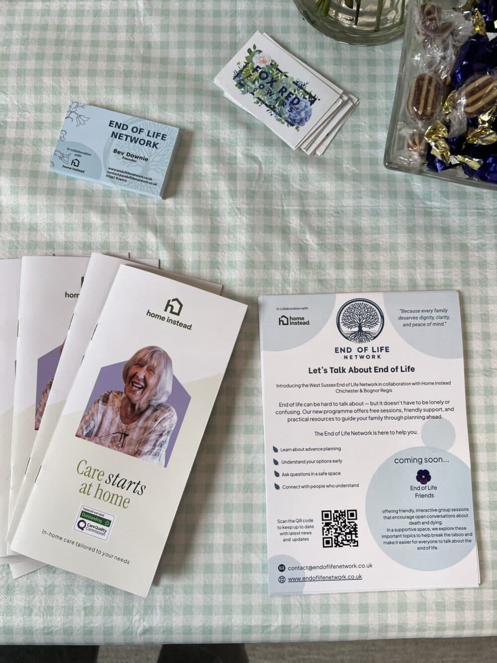 Brochures and flyers about end-of-life care and home care on a checkered tablecloth with business cards and sweets. - Home Instead