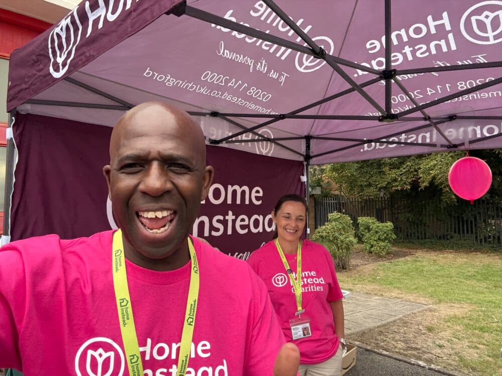 Two people in bright pink "Home Instead" shirts smile under a branded canopy at an outdoor event. - Home Instead
