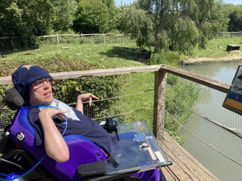 A man in a wheelchair enjoys a sunny day by an animal enclosure with lush greenery and a small pond. - Home Instead