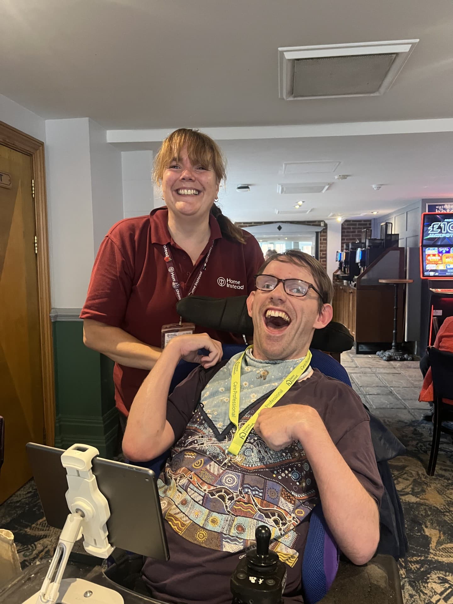 A woman and a man in a wheelchair smile together in a café, with a tablet and drink on the table. - Home Instead