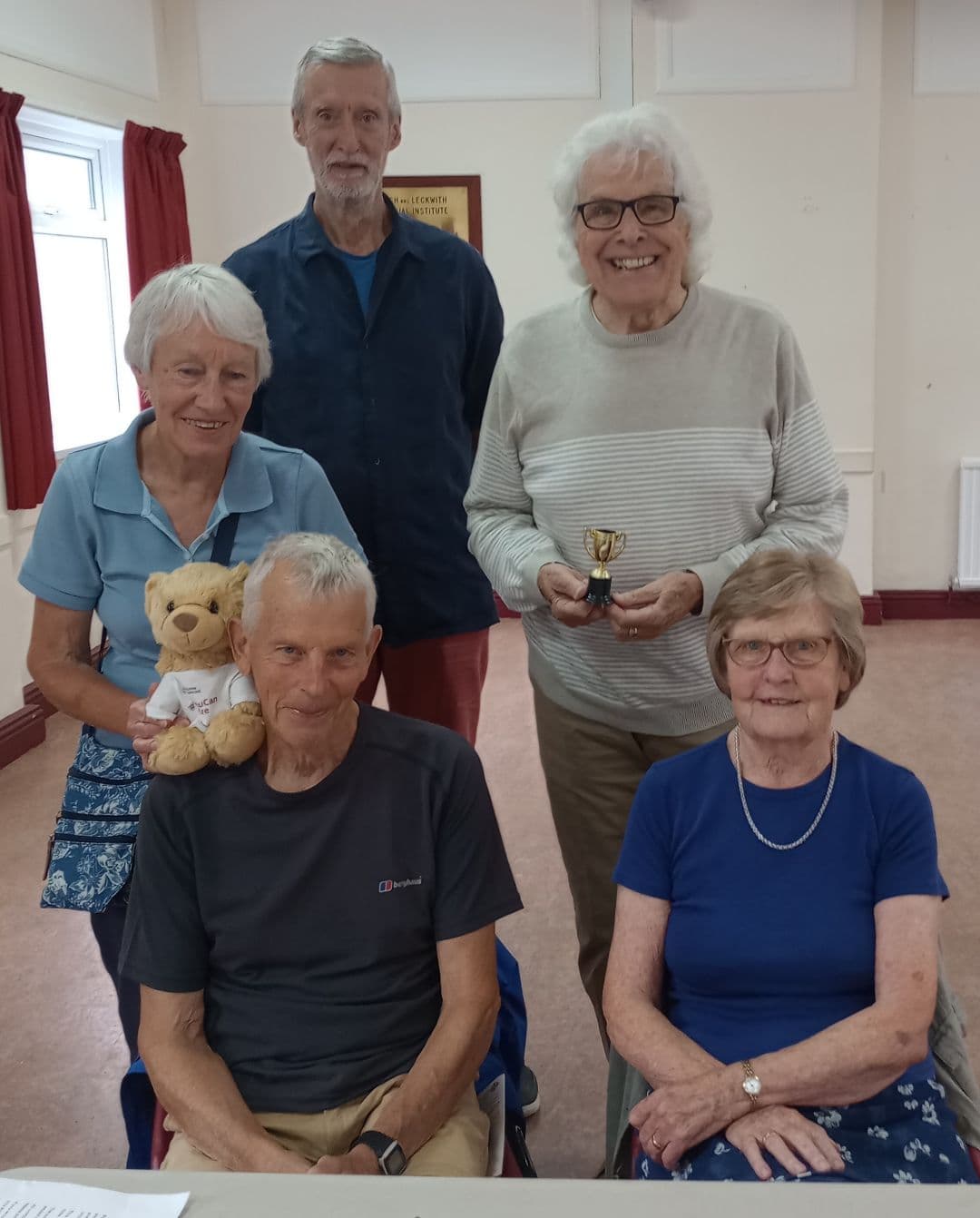 Five smiling older adults, one holding a teddy bear and another holding a small trophy, pose indoors together. - Home Instead
