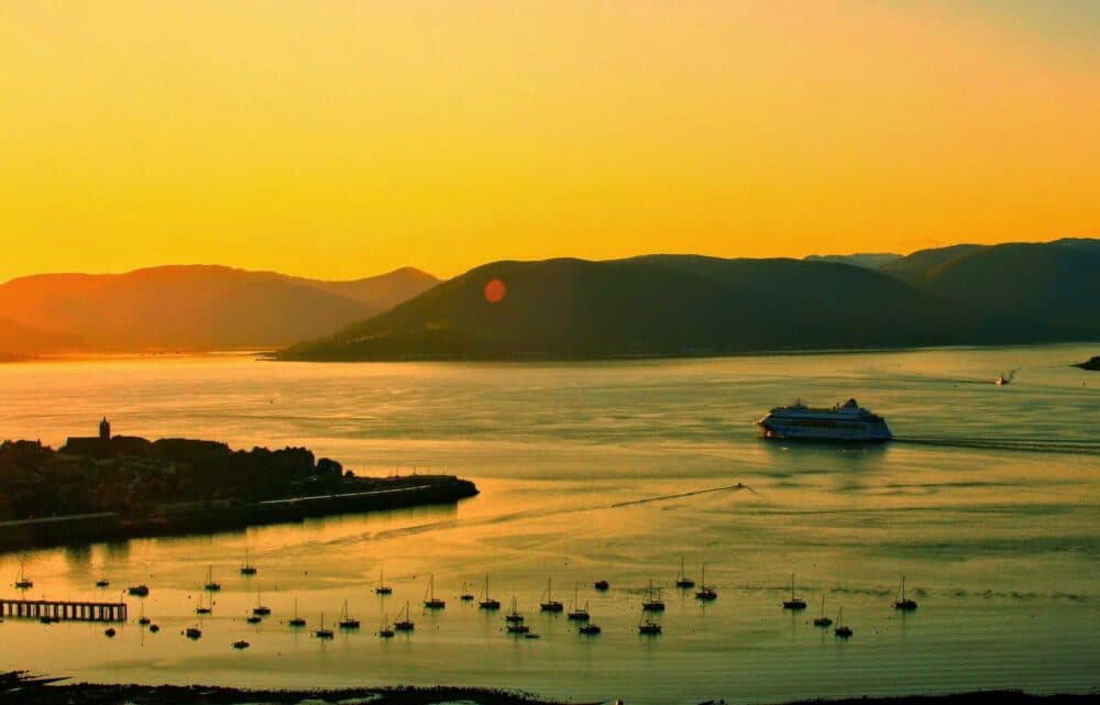 Cruise ship and boats on a calm bay at sunset, with hills and golden sky in the background. - Home Instead