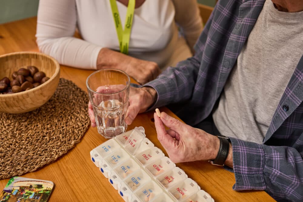 Older adult holding a pill and glass of water near a weekly pill organizer, with a caregiver nearby. - Home Instead