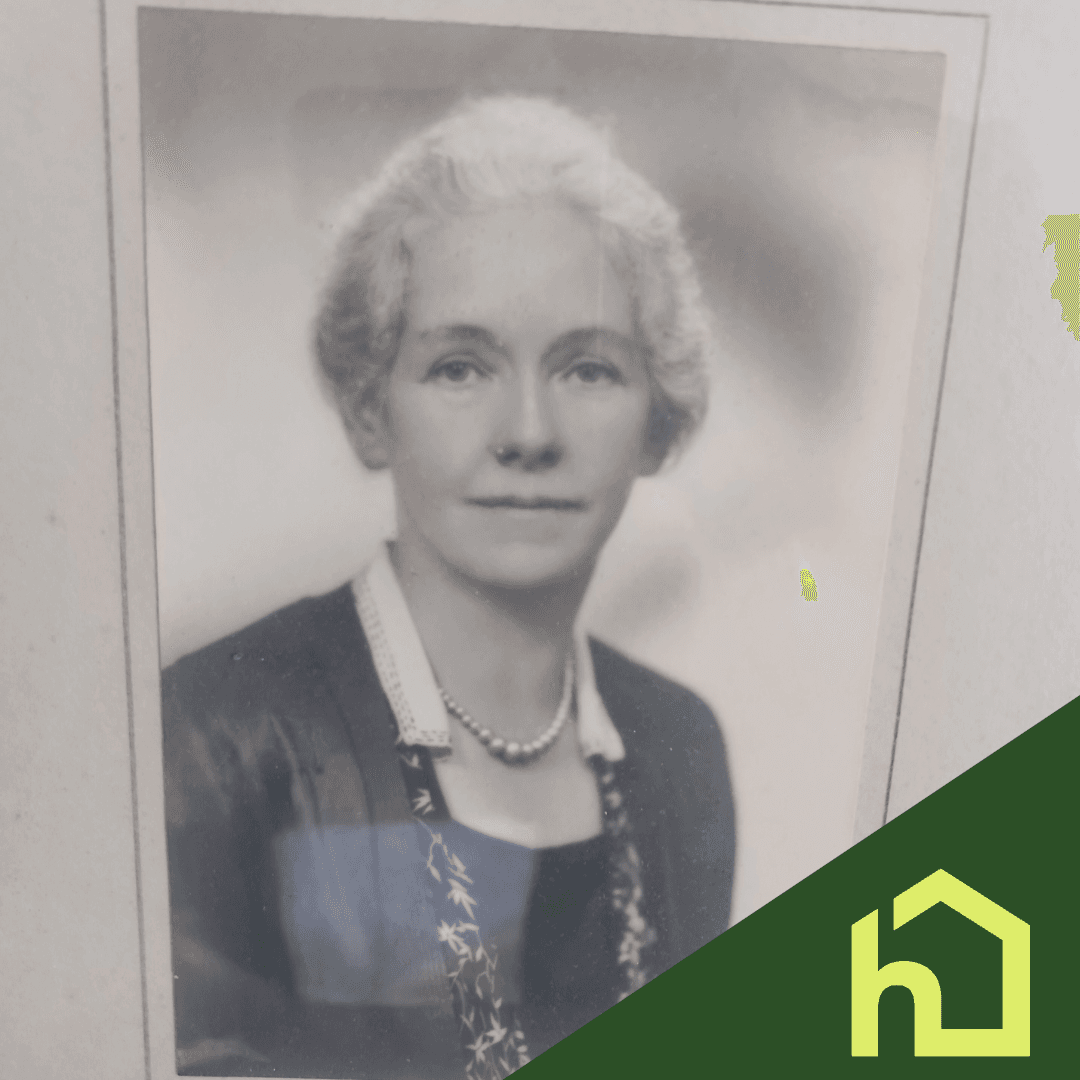 Black and white portrait of an older woman with light hair, wearing a necklace and dark clothing. Green logo overlay. - Home Instead