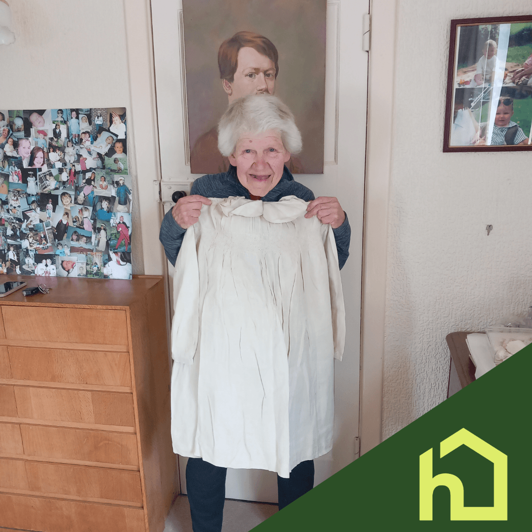 Elderly woman smiling, holding a white dress, standing in a room with family photos and a portrait behind her. - Home Instead