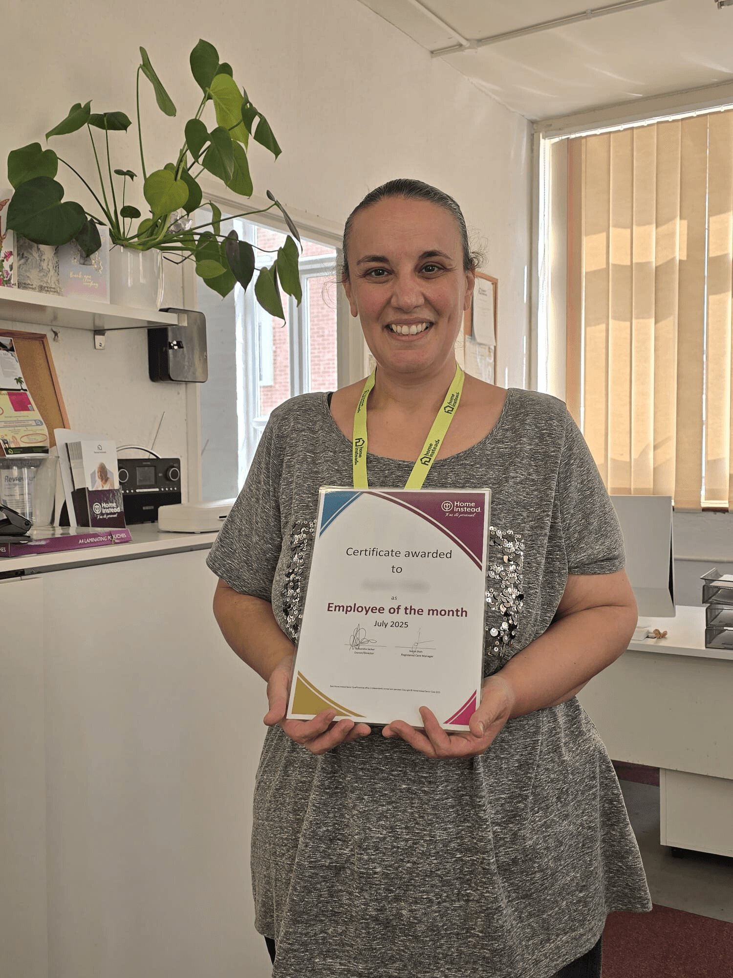 Smiling woman holding an "Employee of the Month" certificate in a bright office space. - Home Instead